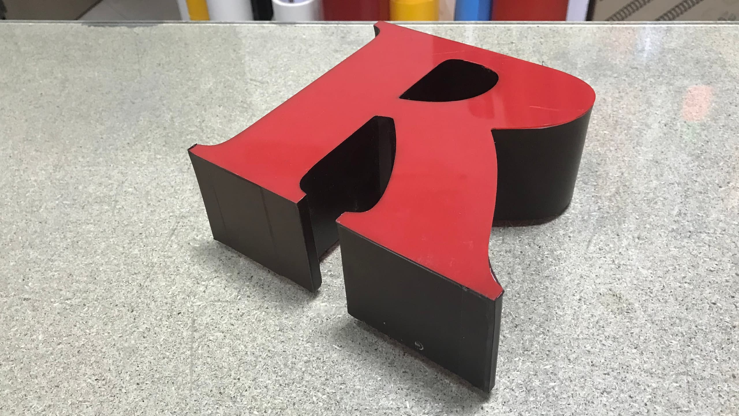 Единична несветеща 3D буква "R" с червено лице от плексиглас и черни страници, поставена на работна маса.