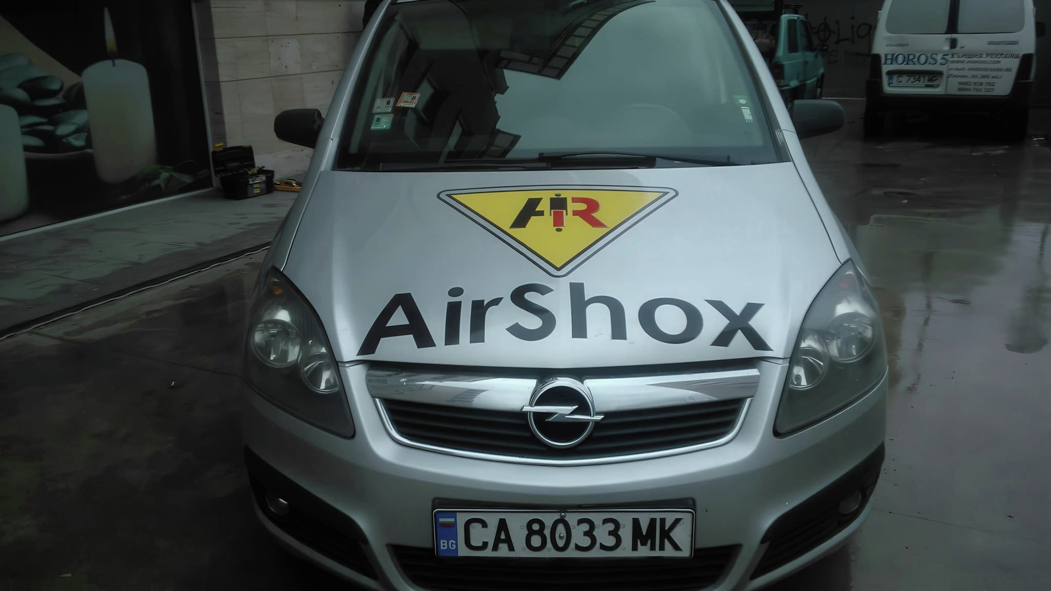Сребрист автомобил Opel с брандиране на предния капак с логото и надписа AirShox, заснет пред сграда с лого на ХОРОС 5 на заден план.