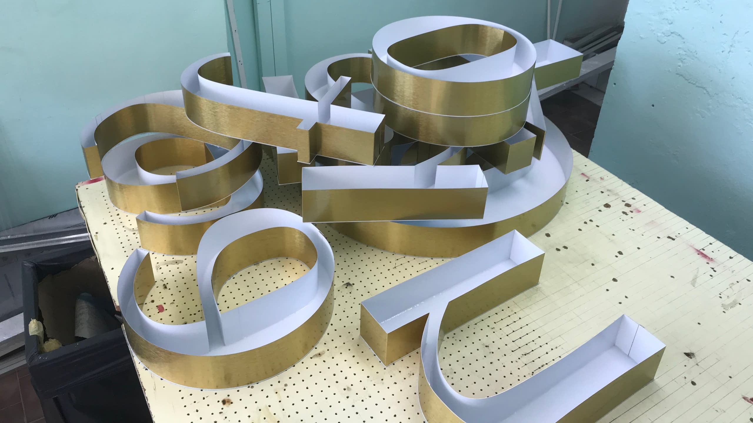 Заготовки за обемни букви - машинно огънати алуминиеви кантове със златно покритие, подредени в рекламна работилница.