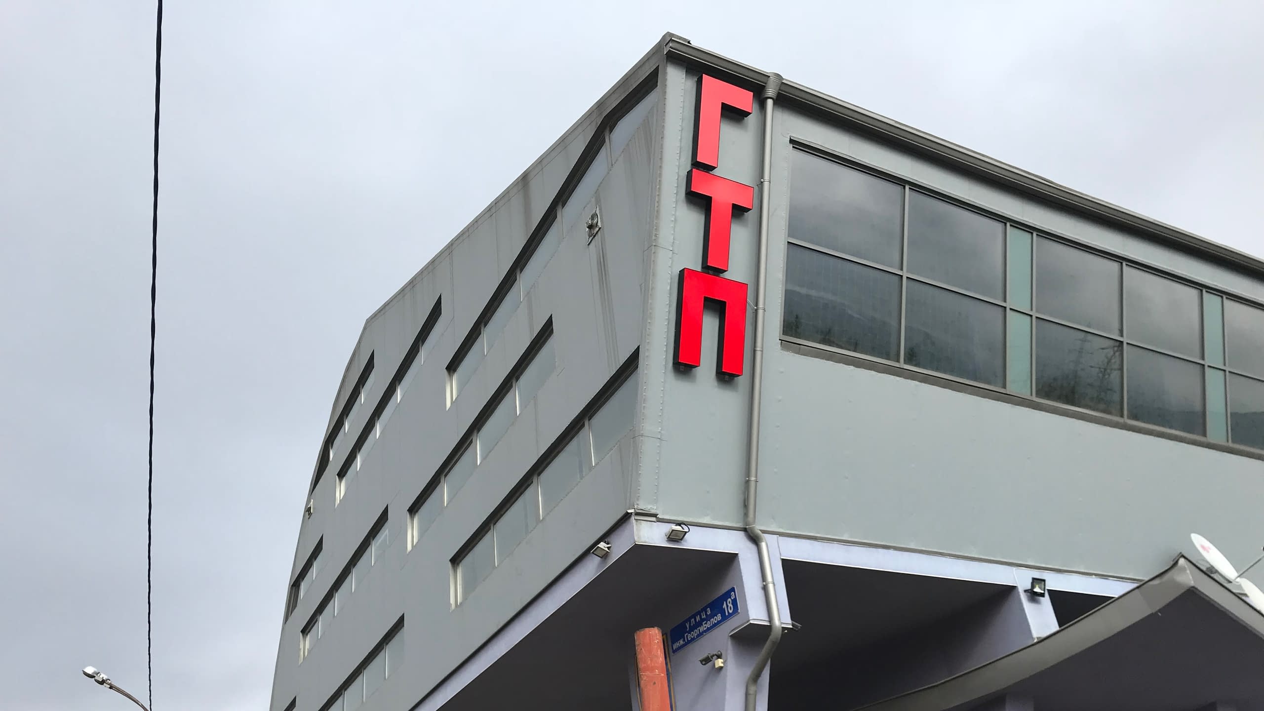 Вертикална рекламна табела от червени обемни букви "ГТП", монтирана на ъгъла на сива сграда.