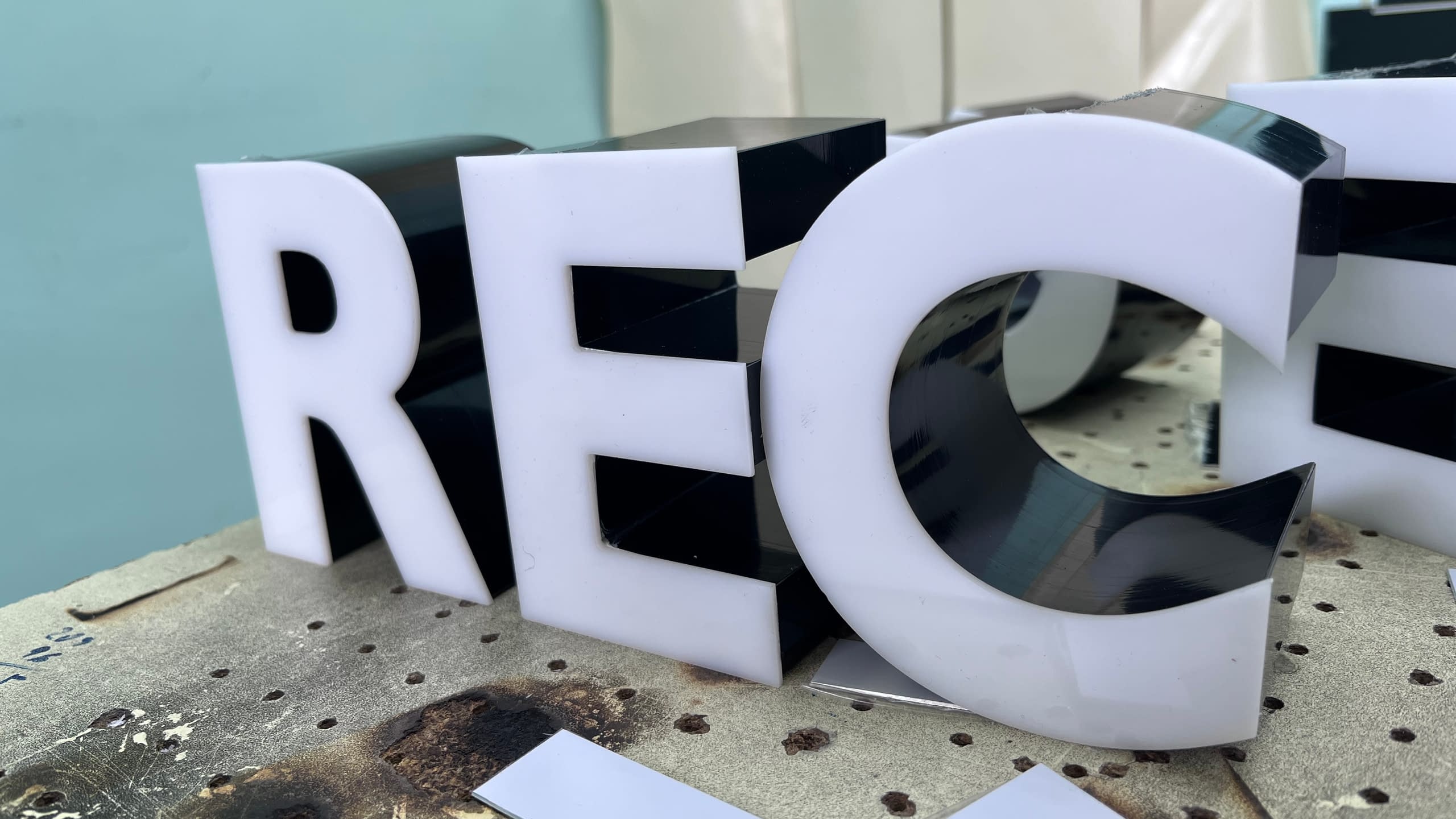 Близък план на обемни букви "REC" с бяло лице от плексиглас и черни гланцови страници, в процес на изработка.