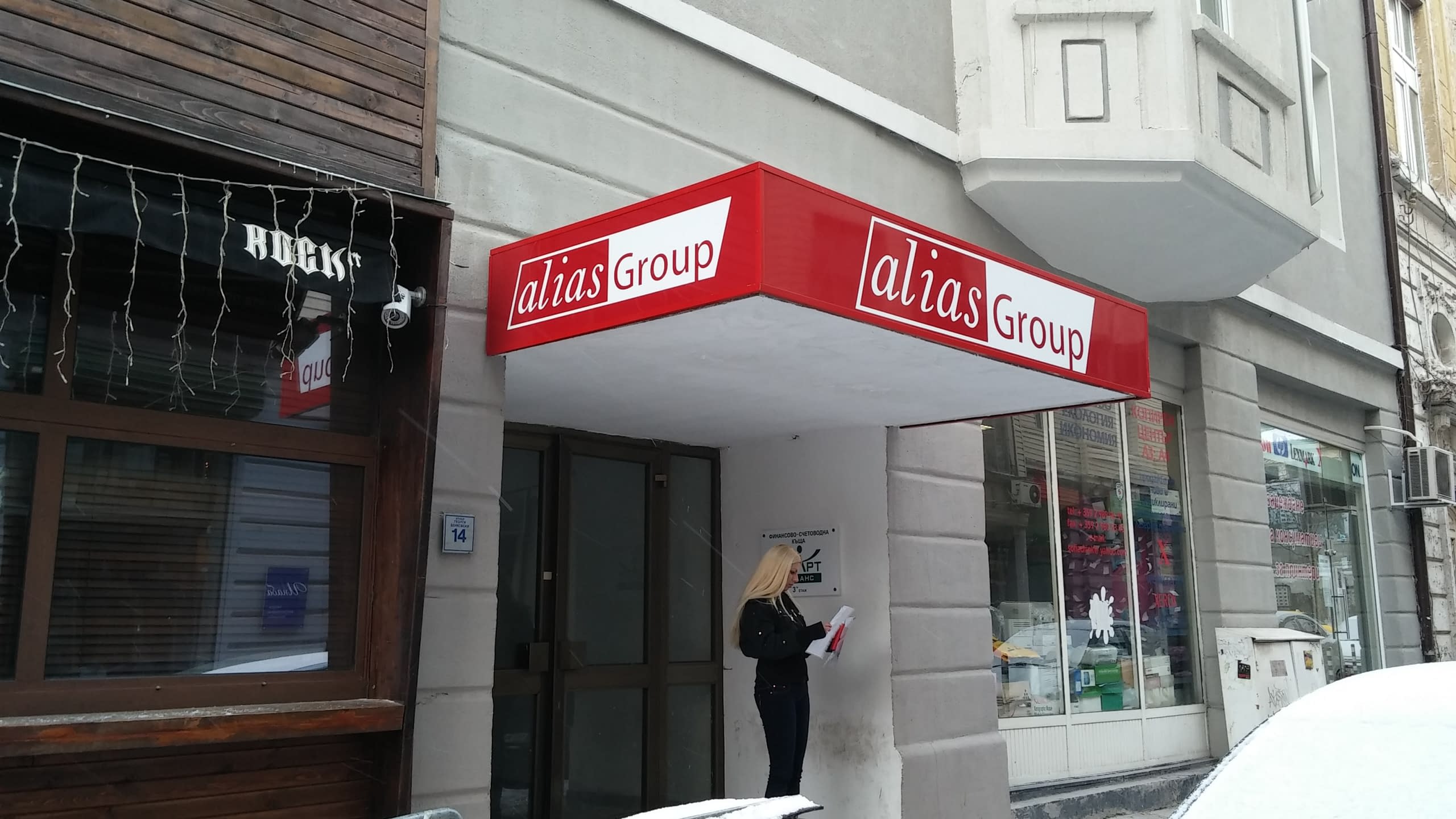 Червена правоъгълна табела с бял надпис "alias Group", монтирана над стъклена врата на сграда.