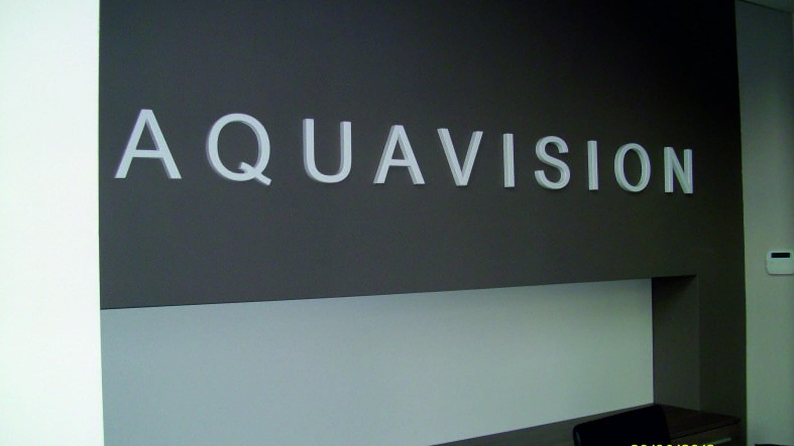 Бели обемни букви с надпис AQUAVISION, монтирани на тъмносива стена в офис.