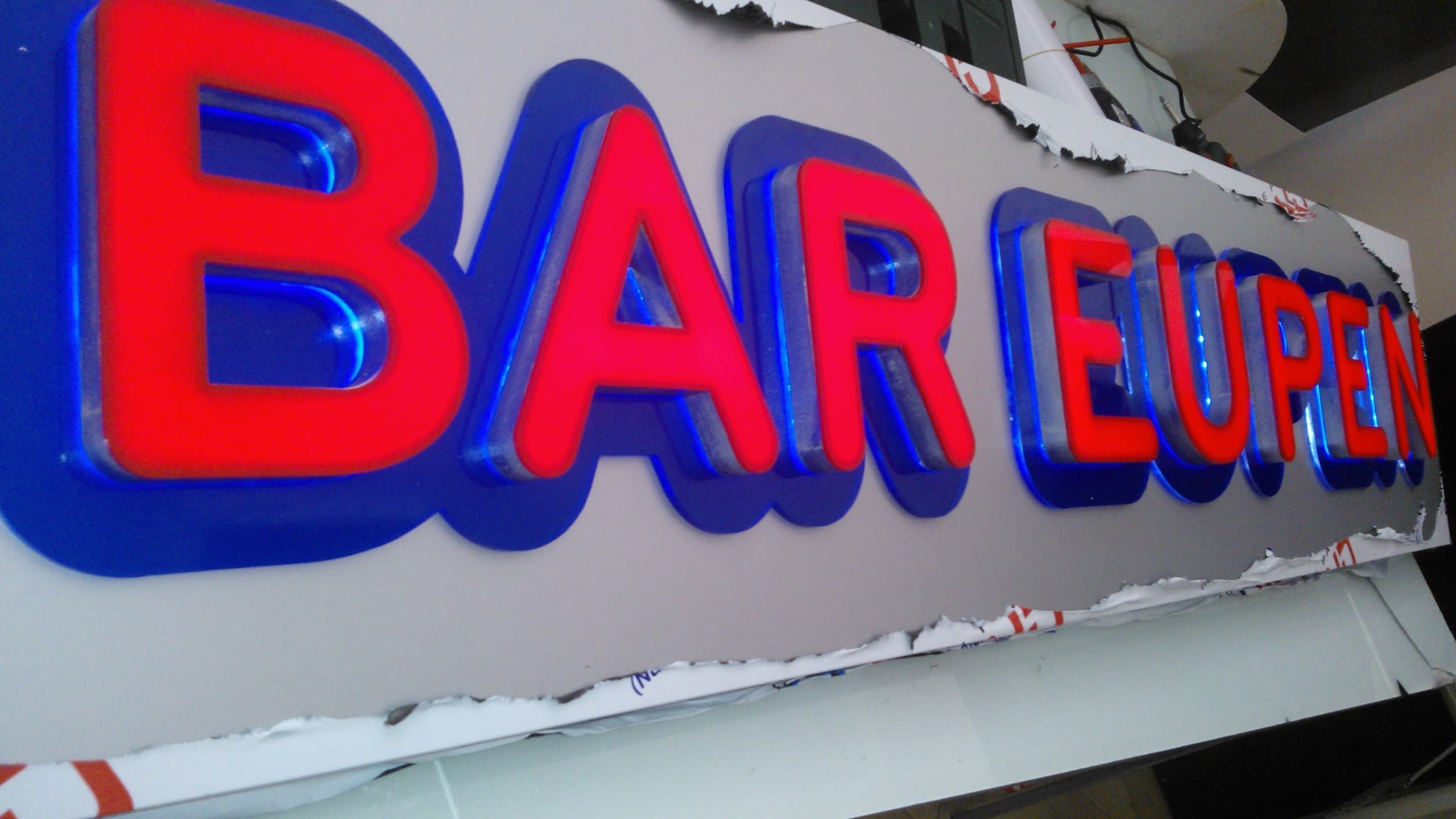 Светещ надпис „BAR EUPEN“, монтиран върху тухлена стена на сграда и сниман отдолу.