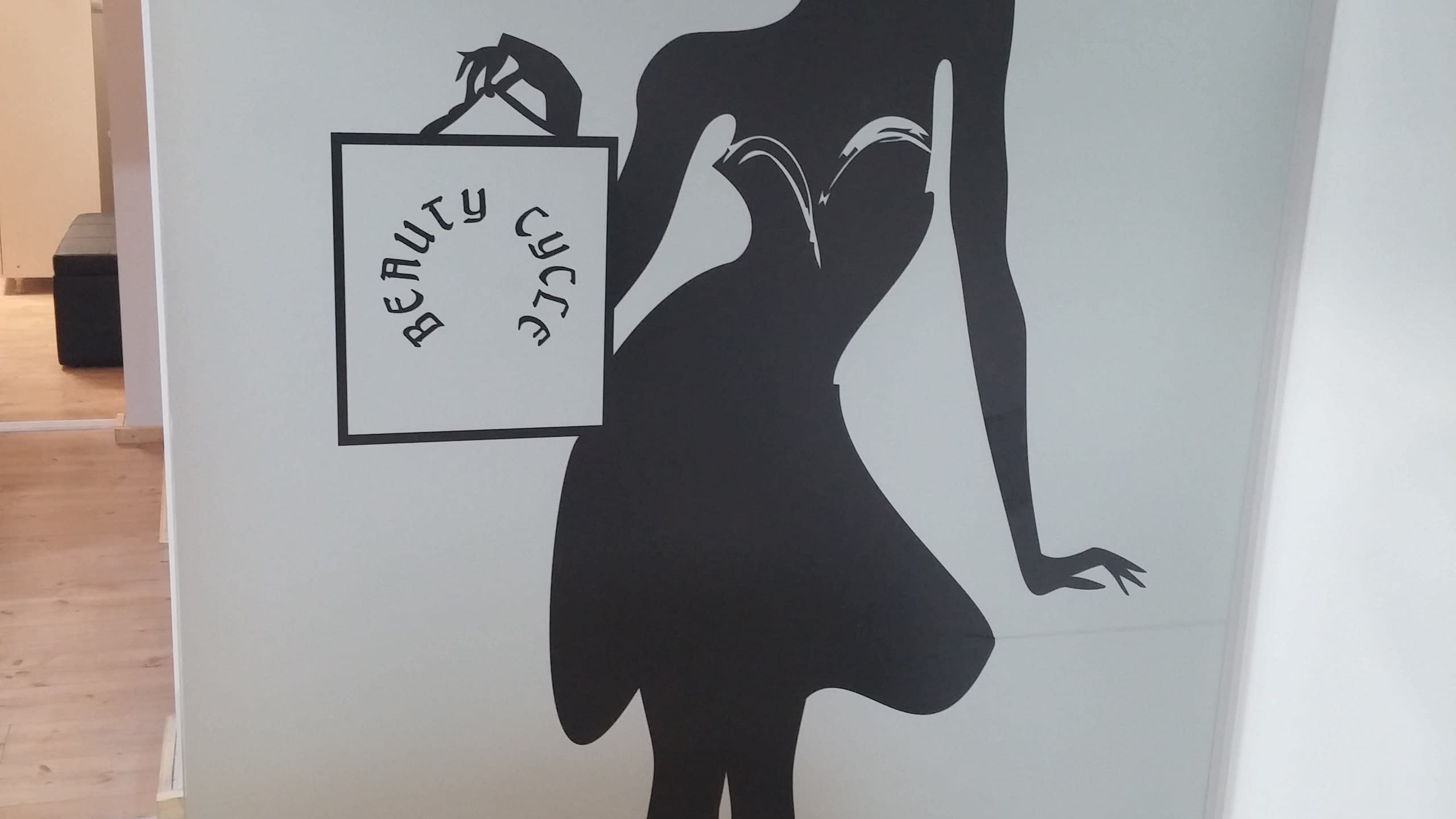 Черна графика на жена с голяма шапка и пазарска чанта с надпис, апликирана върху светъл панел в интериорно пространство.