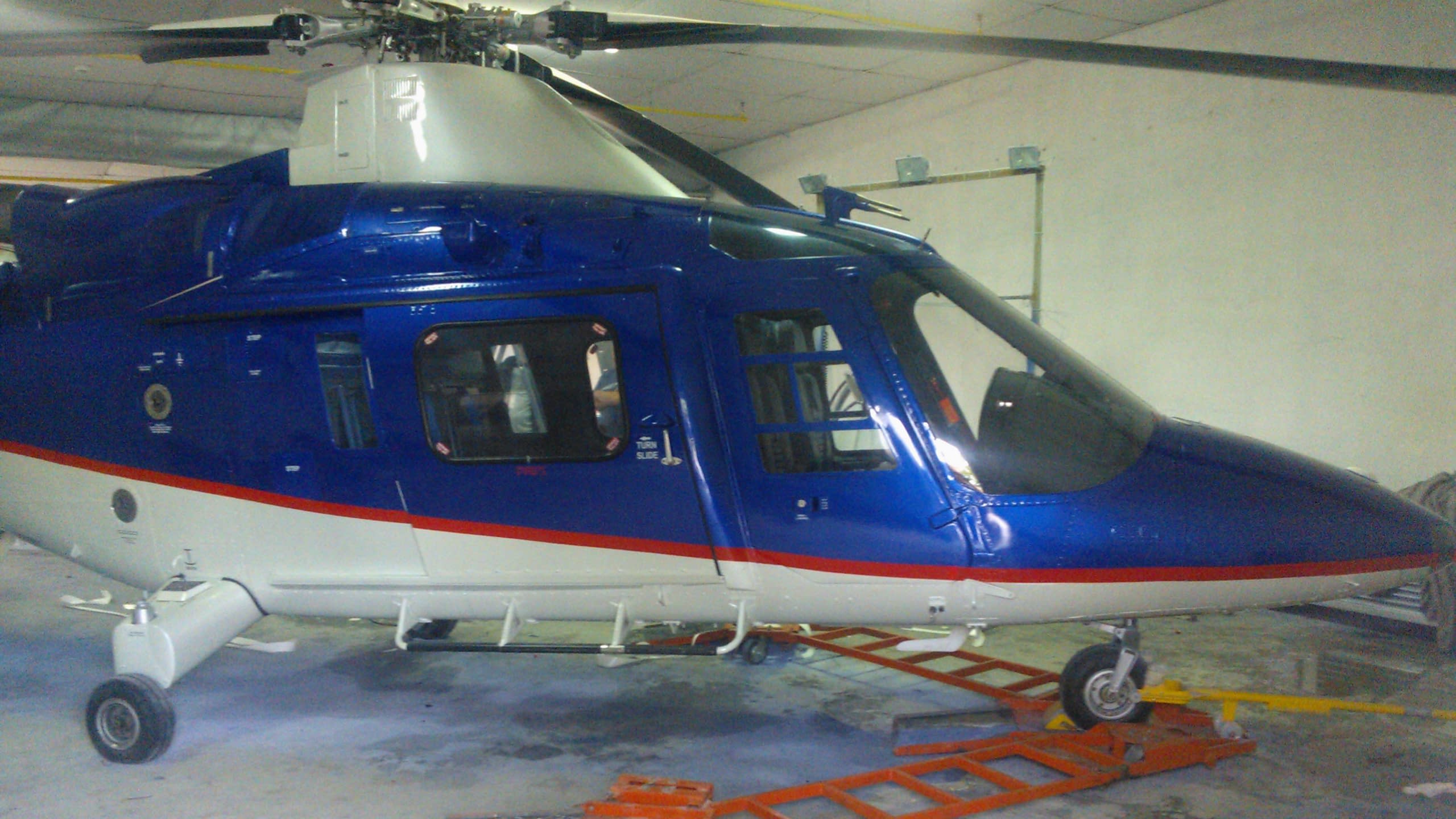 Хеликоптер в синьо и бяло, разположен върху оранжева платформа за преместване в хангар.