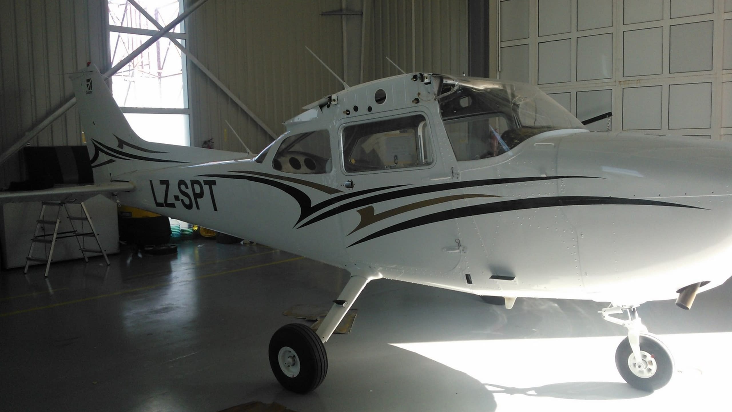 Малък бял самолет с регистрация LZ-SPT и декоративни черни и златни ленти, паркиран в хангар.