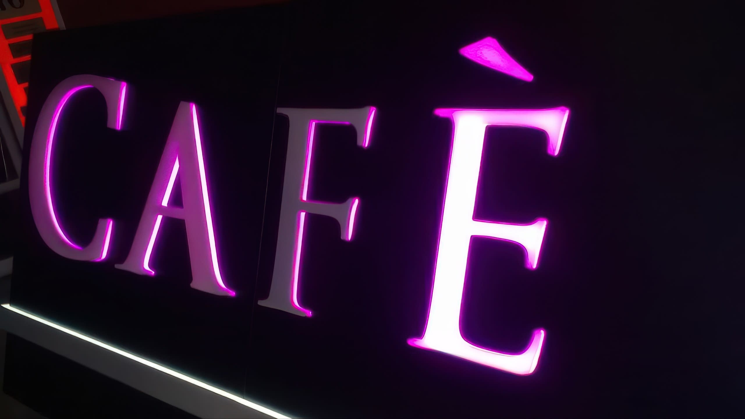 Близък план на надпис „CAFÈ“, чиито букви светят с наситена розова светлина на тъмен фон.