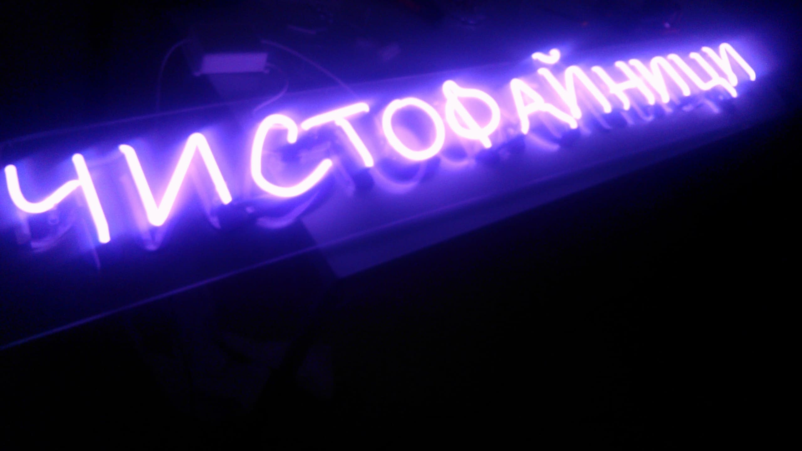 Светещ надпис на кирилица с лилав цвят, заснет в пълен мрак, за да се подчертае сиянието на извитите тръби.