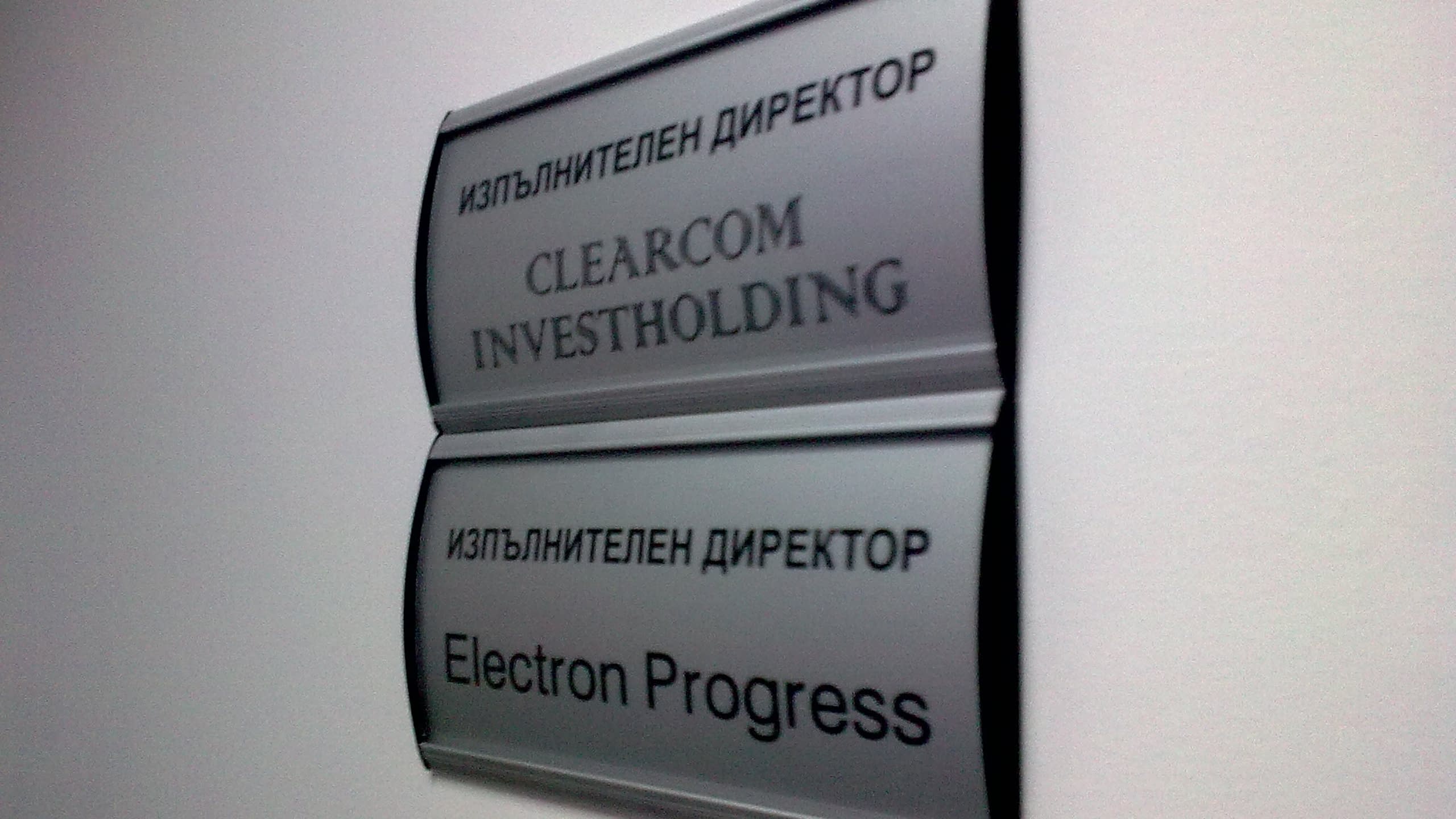 Две сиви табелки за врата, монтирани една над друга на стена, с надписи за длъжност и име на фирма.
