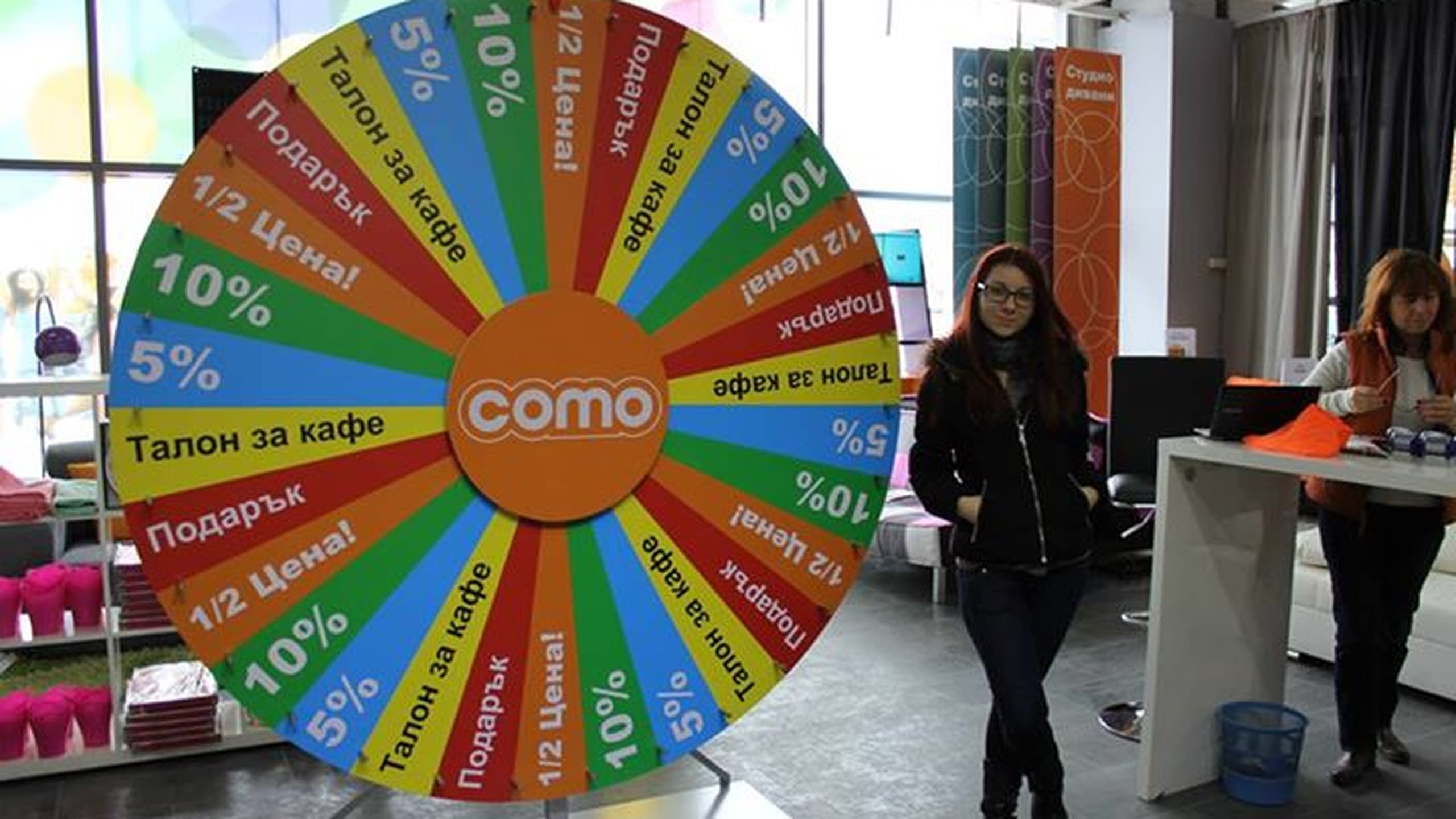 Голямо цветно колело с надписи за награди, поставено в интериора на магазин, с посетители на заден план.