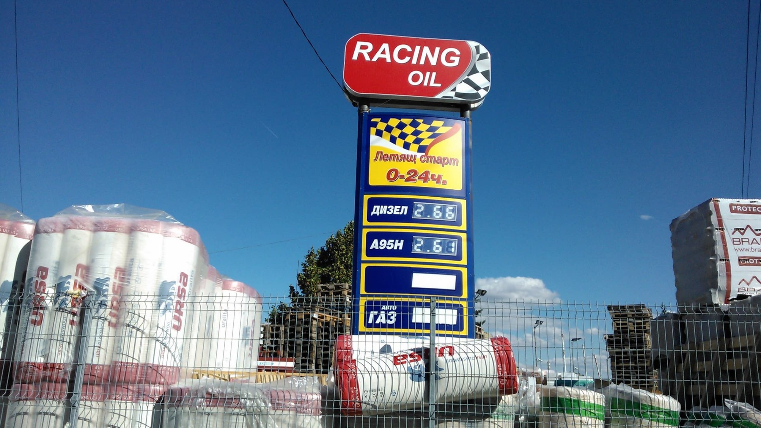 Висок рекламен тотем за бензиностанция с табела "Racing Oil" и светещи панели, показващи цени на горивата.