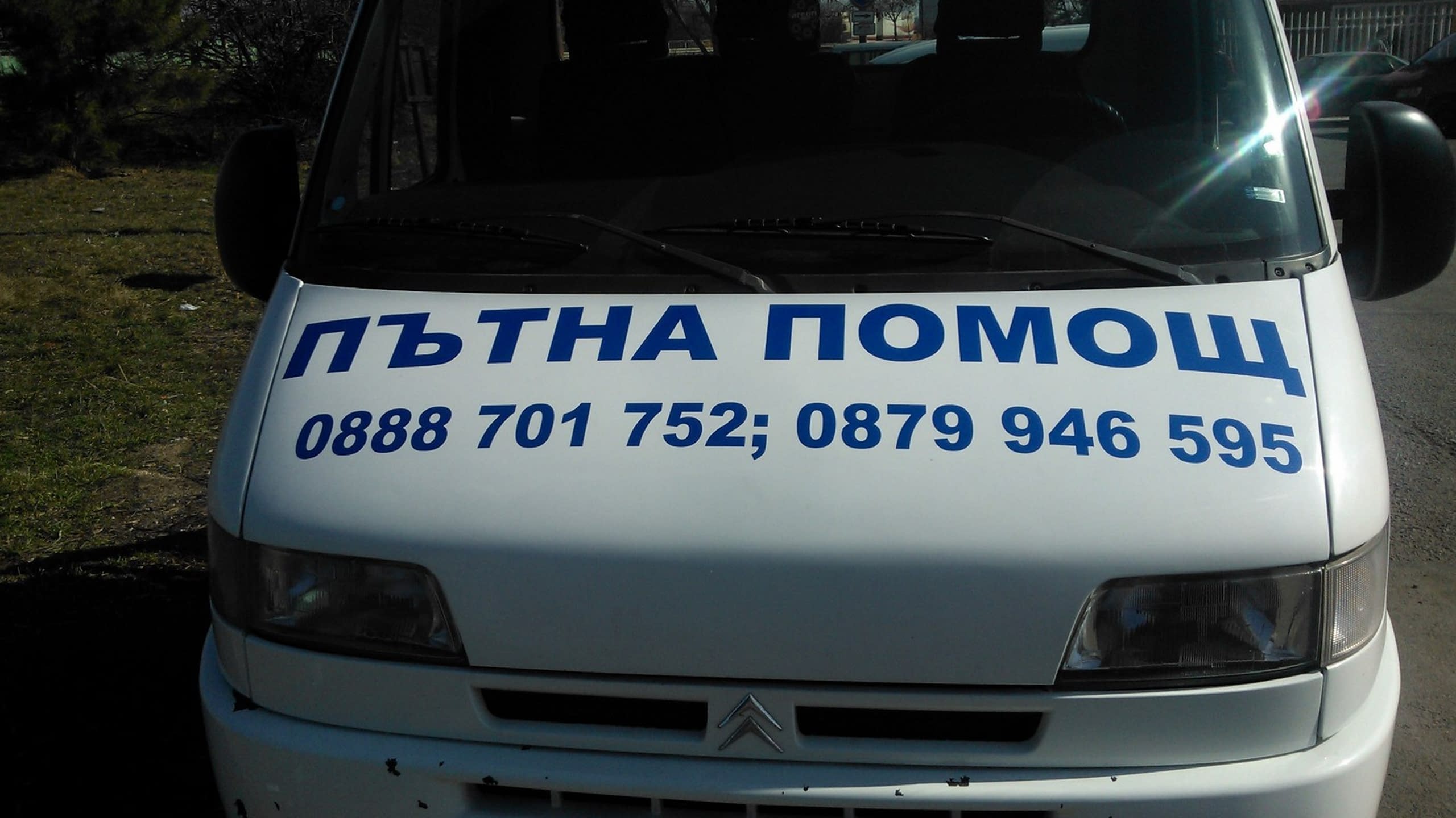 Предница на бял камион с надпис „Пътна помощ“ и телефонни номера, изписани със сини букви на капака.