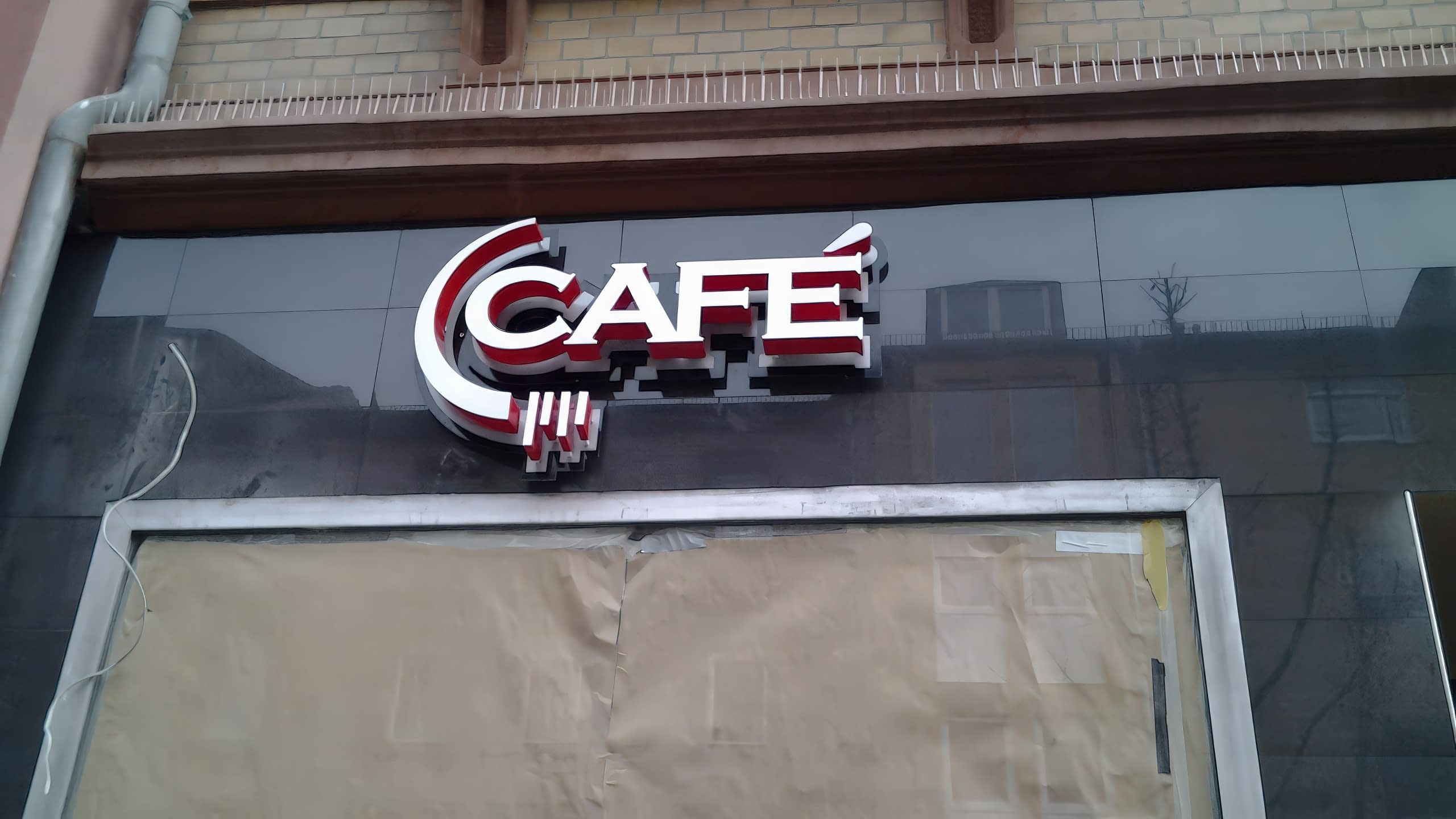 Монтиран надпис „Café“ на черна лъскава фасада над прозорец, покрит с хартия.