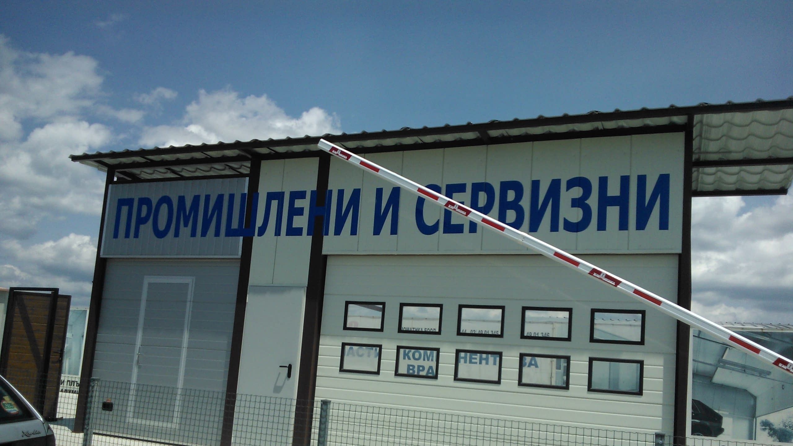 Фасада на индустриална сграда със секционна врата и голям надпис със сини букви, частично закрит от вдигната червено-бяла бариера.