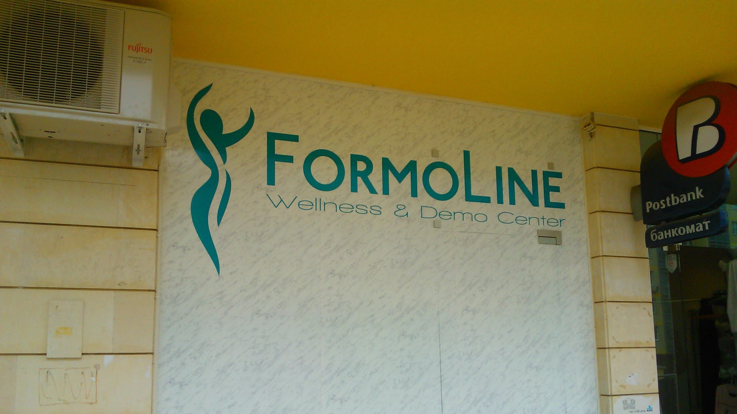 Лого на Formoline, поставено върху релефна стена до входа на сграда, под жълт таван.