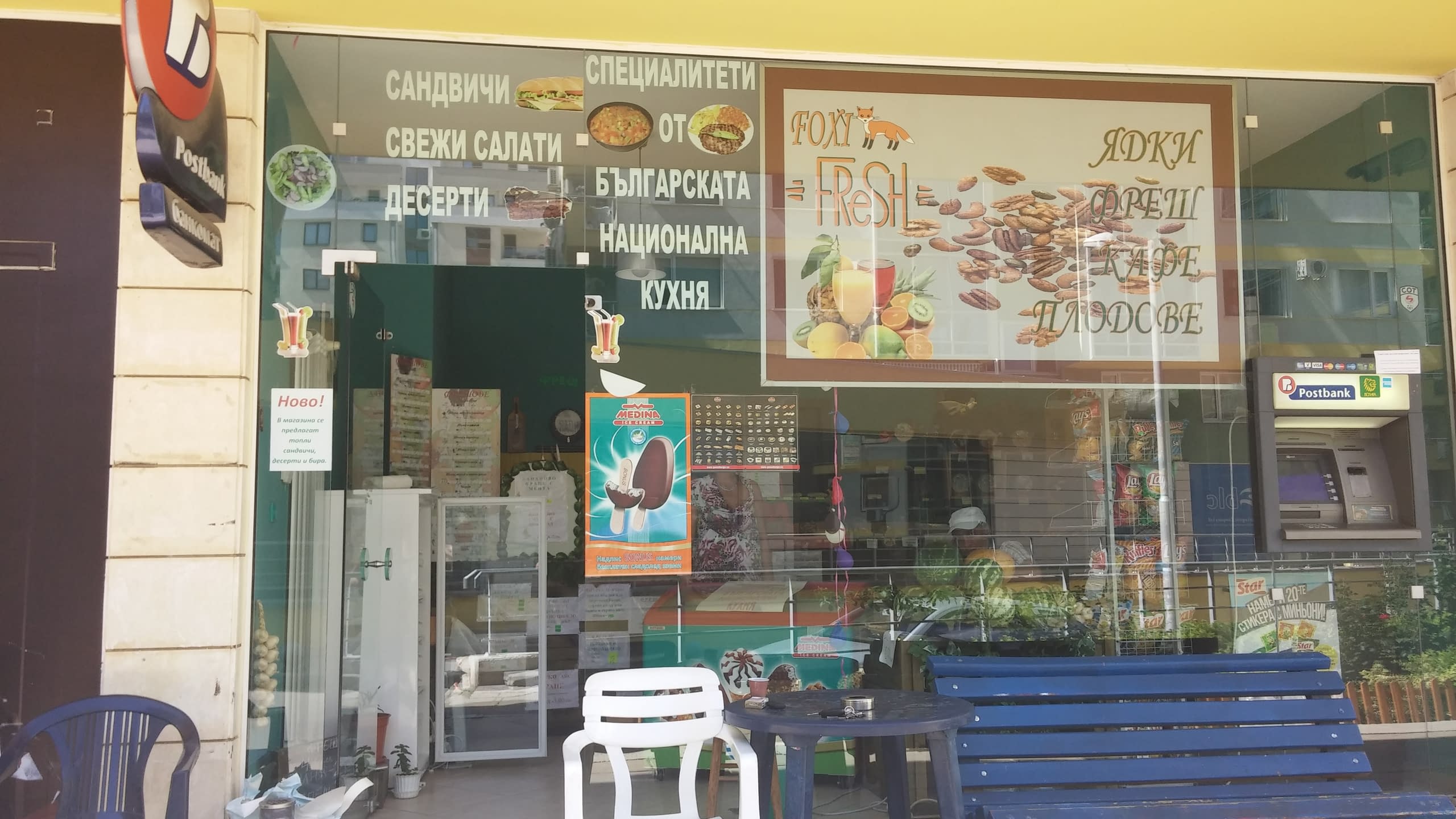 Стъклена витрина на хранителен магазин със стикери, които изброяват предлагани храни като сандвичи, салати и ядки.