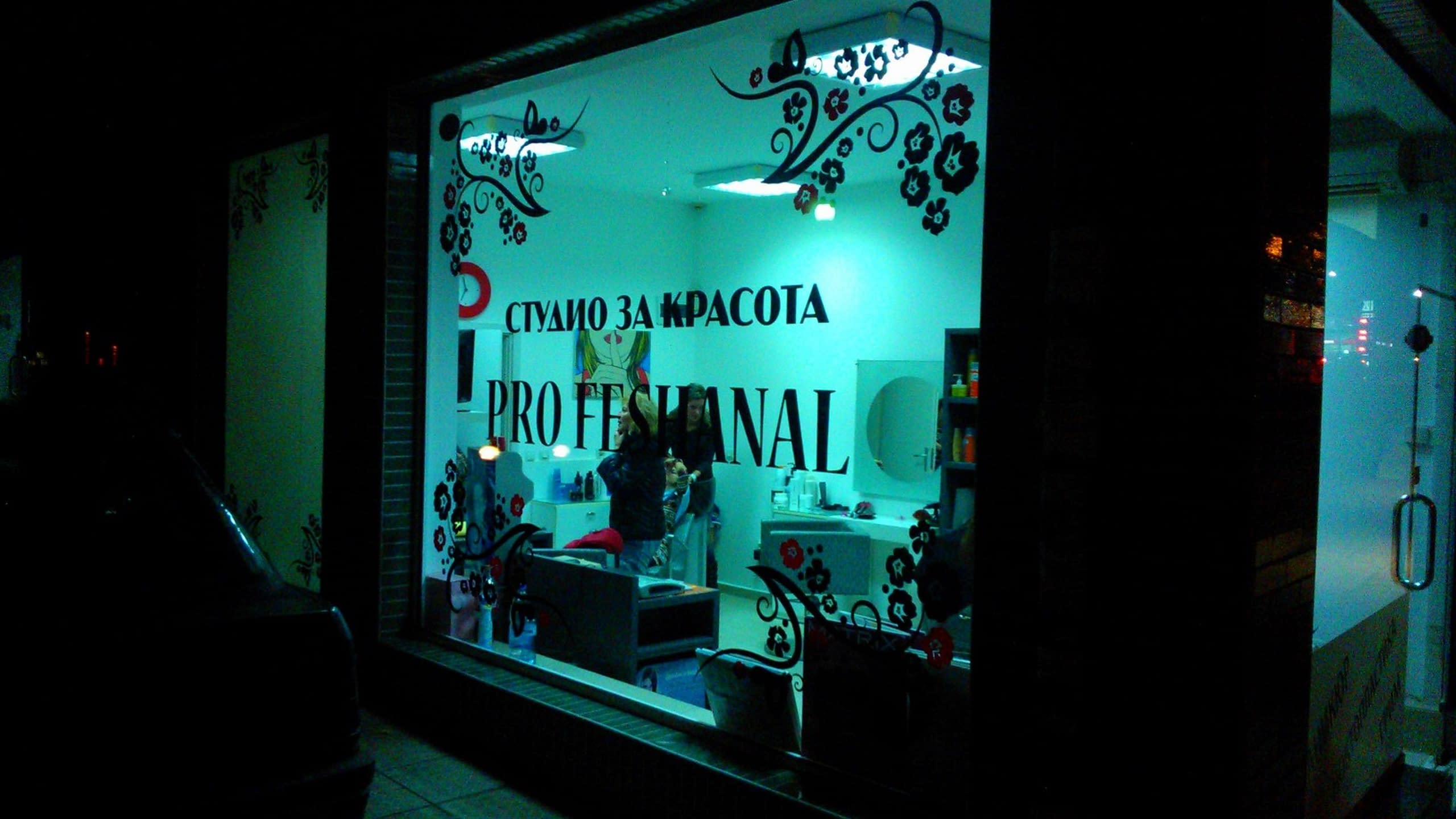 Нощна снимка на осветена витрина на салон за красота, декорирана с черни орнаменти и надписи, през която се вижда интериорът и работещи хора.