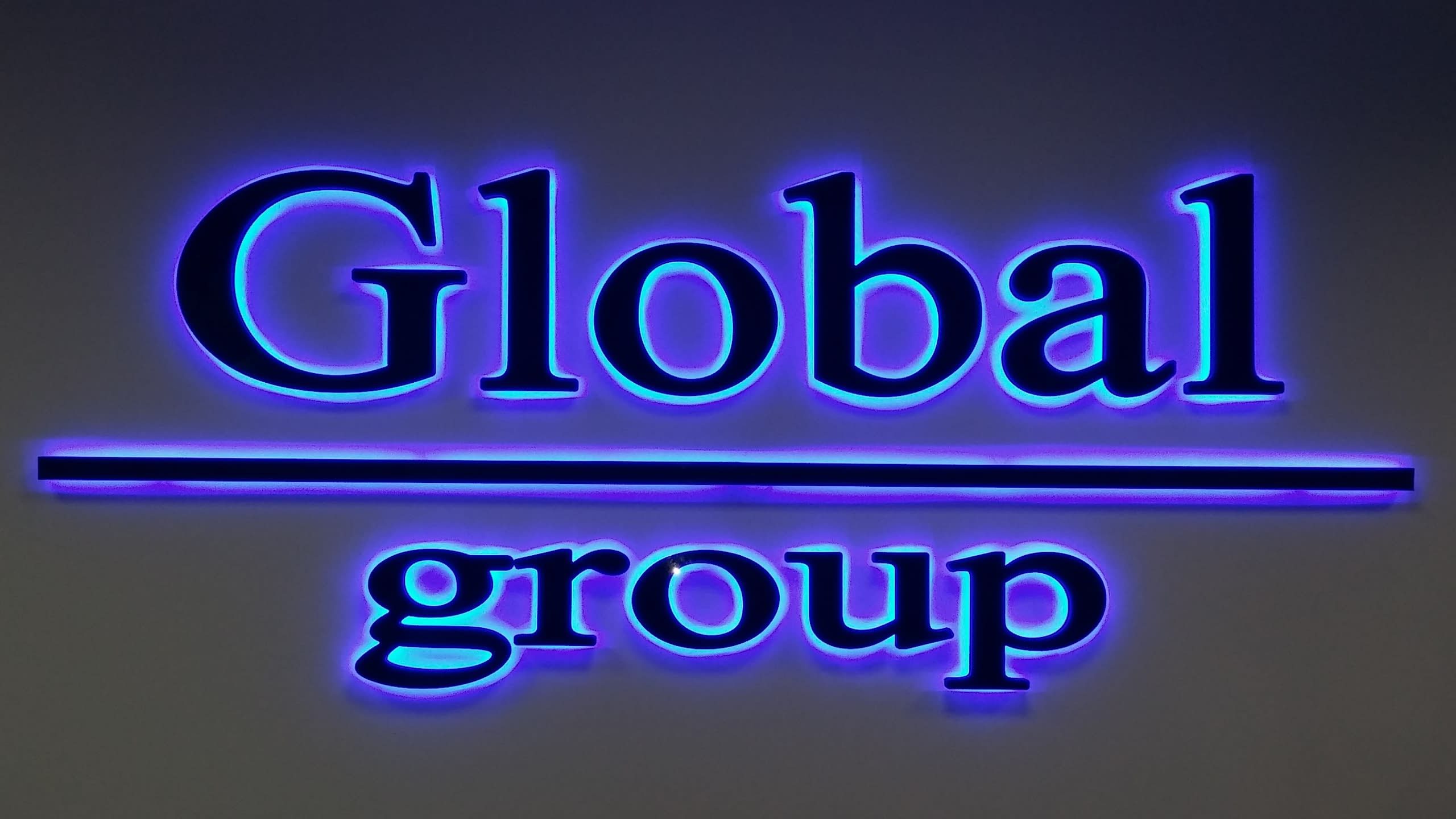 Челен изглед на осветен надпис „Global group“, показващ ореола от синя светлина около тъмните букви.