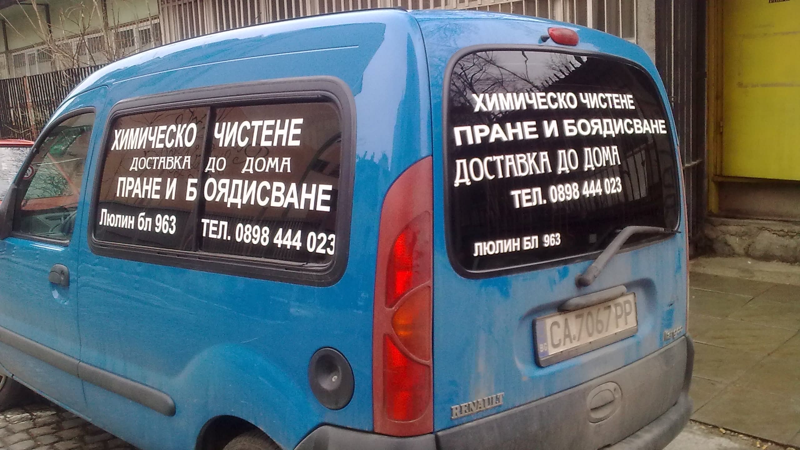 Задната част на син автомобил тип баничарка, чиито странично и задно стъкло са облепени с бели надписи, предоставящи информация за услуги и контакти.