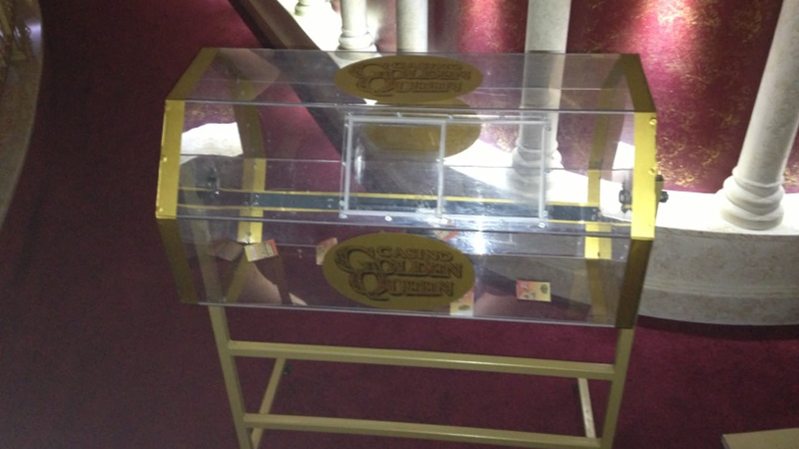 Прозрачен барабан за томбола със златни кантове и лого на "Casino Golden Queen", поставен в интериор с червен фон и колони.