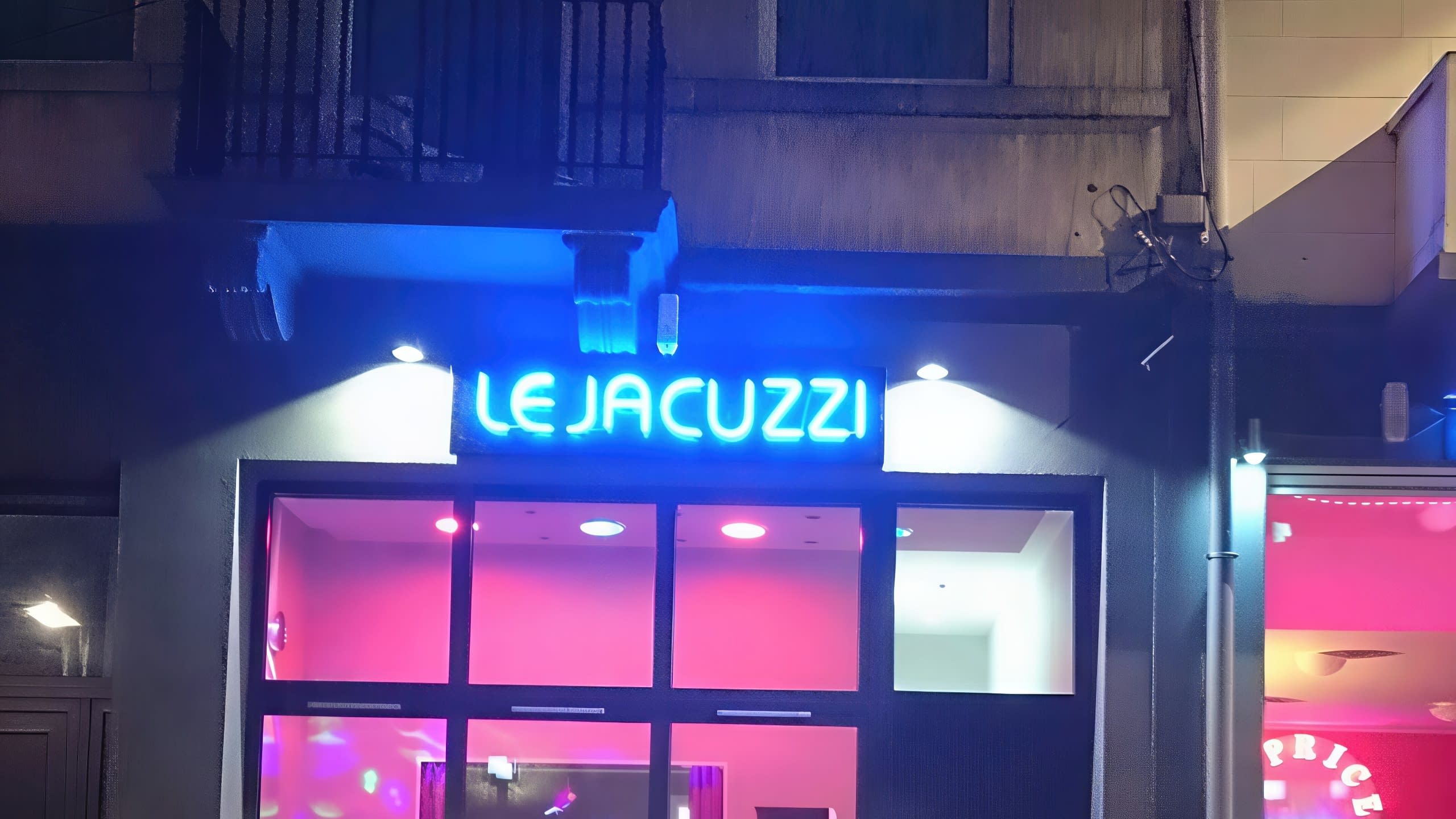 Вход на заведение през нощта, с ярко осветен в синьо надпис LE JACUZZI и розова светлина, идваща от интериора.