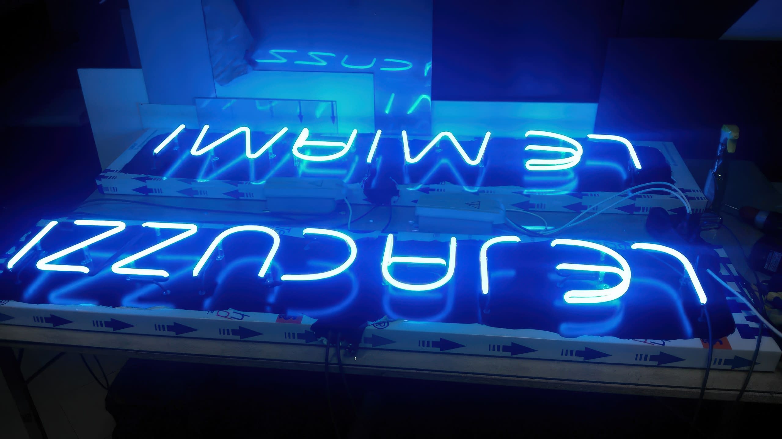 Два светещи в синьо надписа, LE JACUZZI и LE MIAMI, поставени един до друг на работни маси в тъмно помещение.