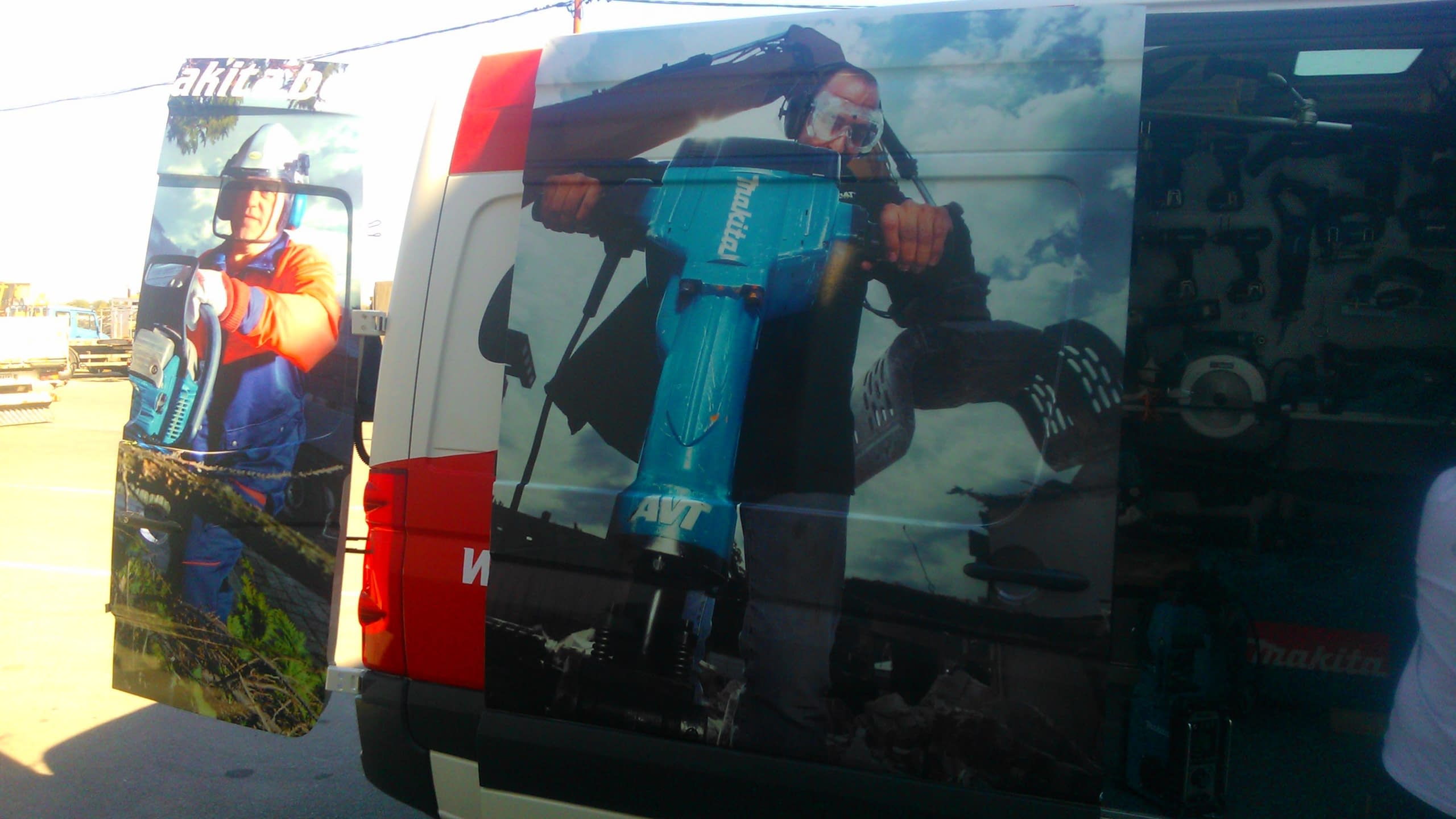 Страничен изглед на брандиран микробус с голямо изображение на човек с къртач и отворена задна врата, която показва интериора с изложени инструменти.