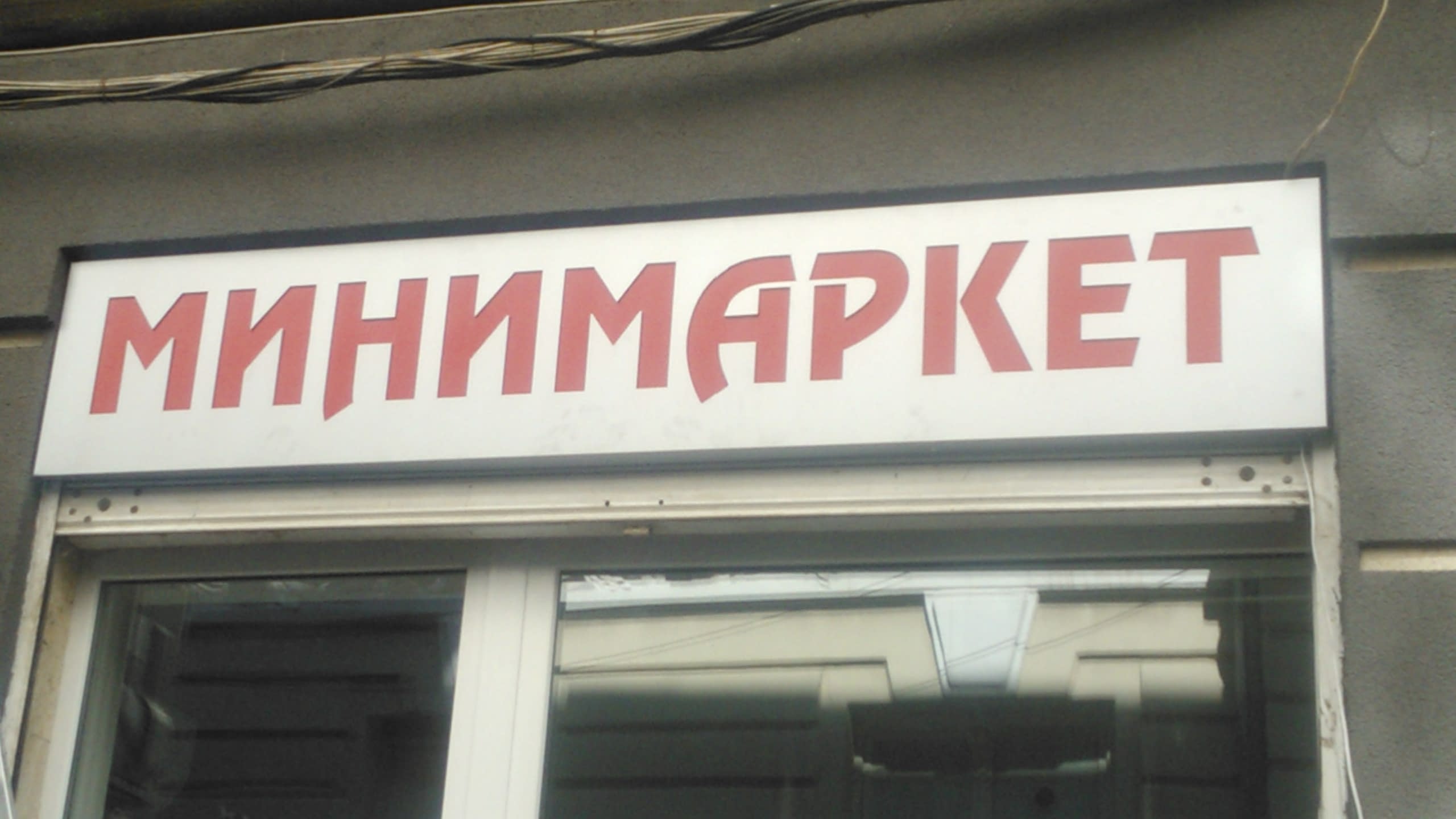 Правоъгълна табела с червен надпис "Минимаркет", монтирана на фасада над магазин.