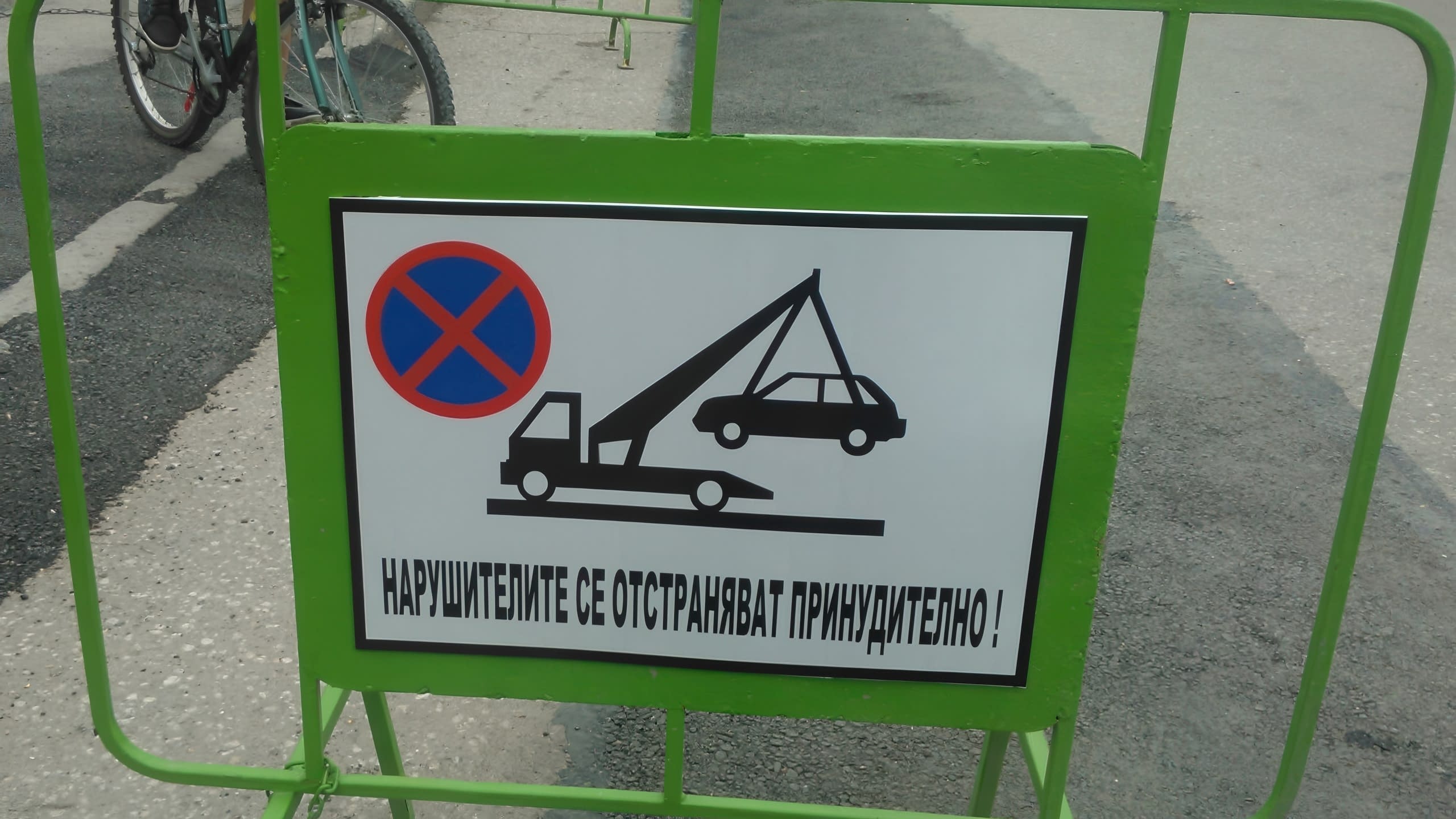 Предупредителна табела за принудително отстраняване на автомобили, монтирана на зелена подвижна ограда.