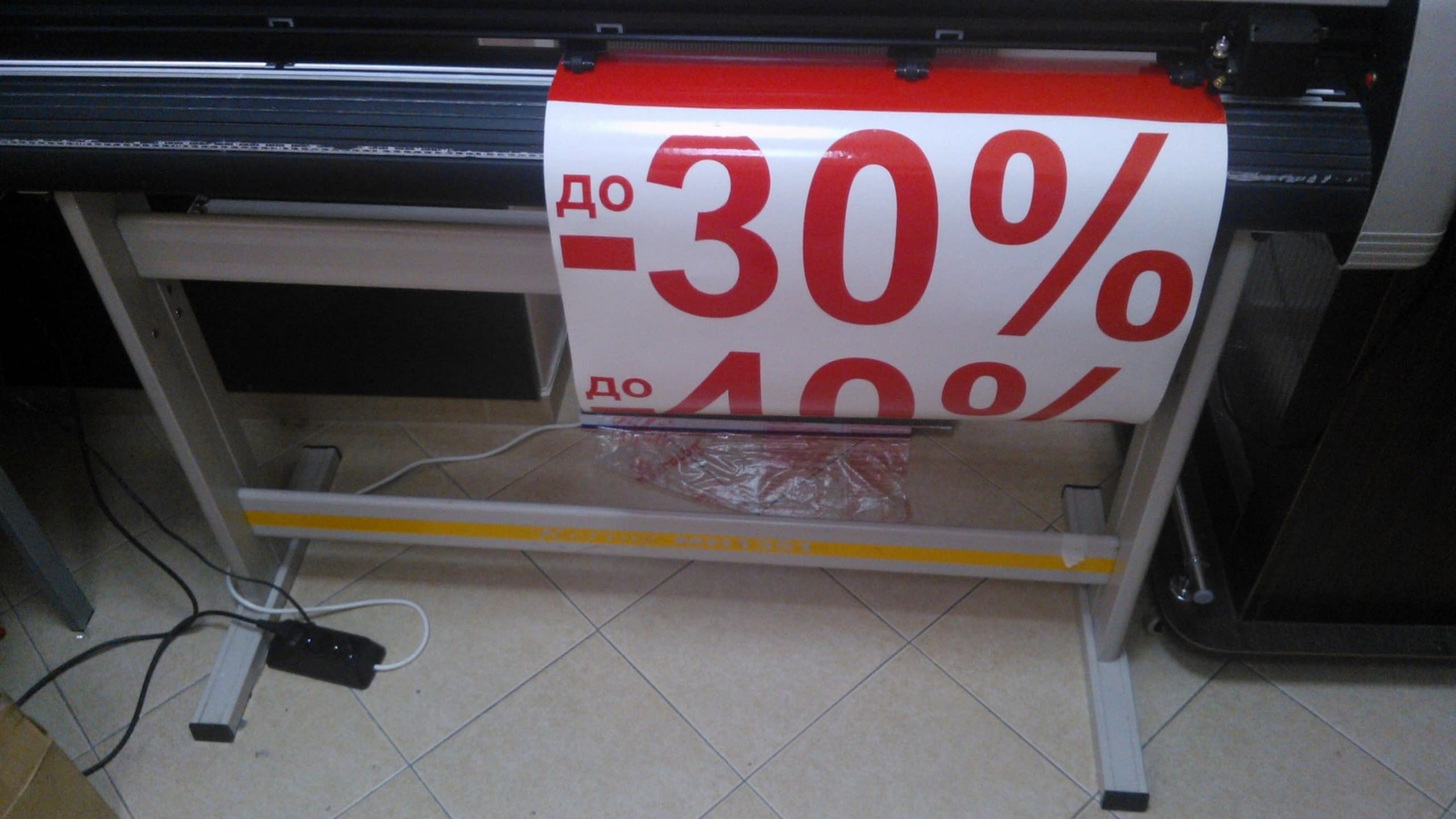 Машина с режеща глава обработва ролка бяло фолио, от което изрязва надписи с червени цифри и знаци за процент.