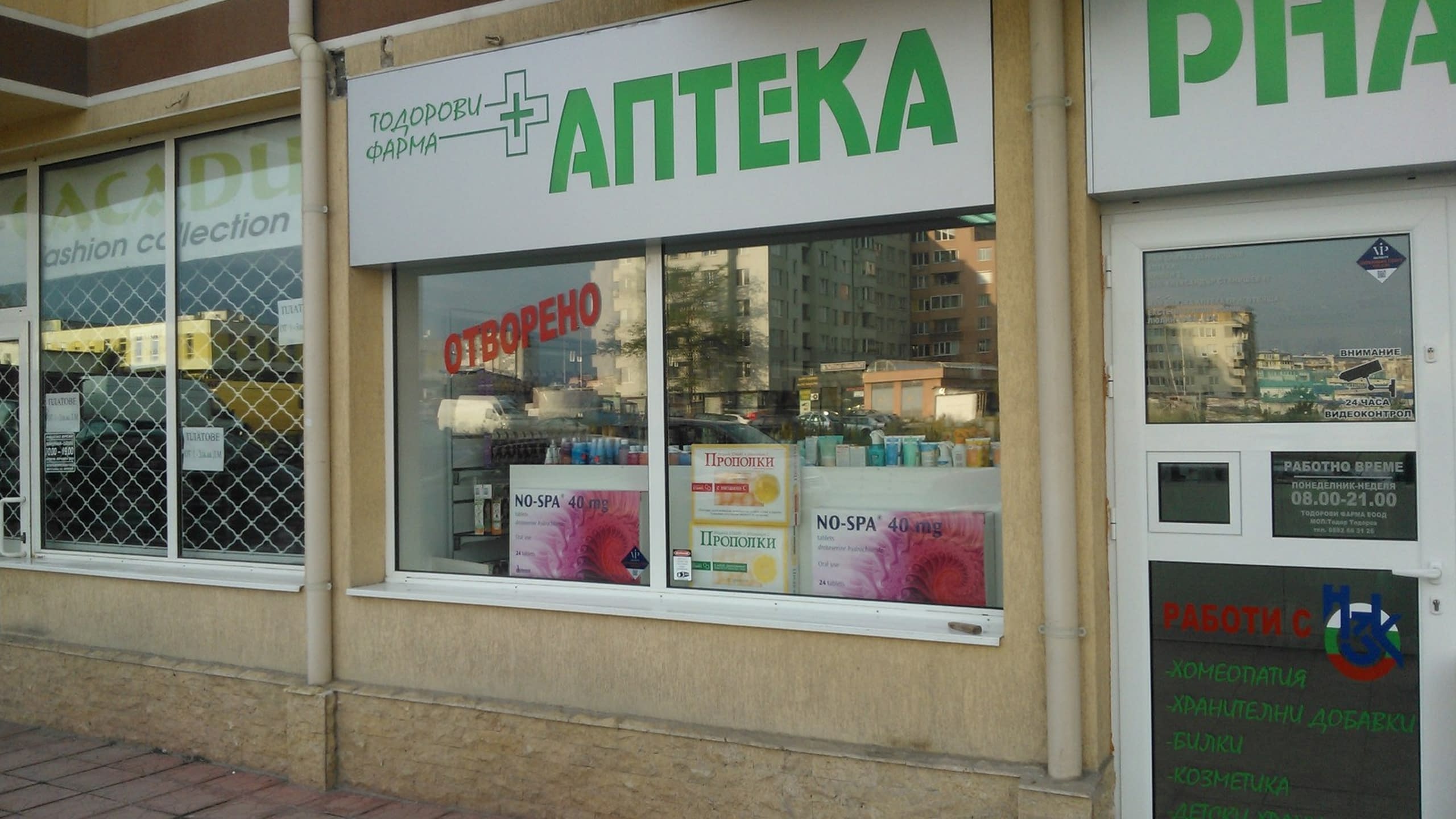 Фасада на аптека с монтирана правоъгълна бяла табела, на която със зелени букви е изписано „АПТЕКА“ и името на фирмата.