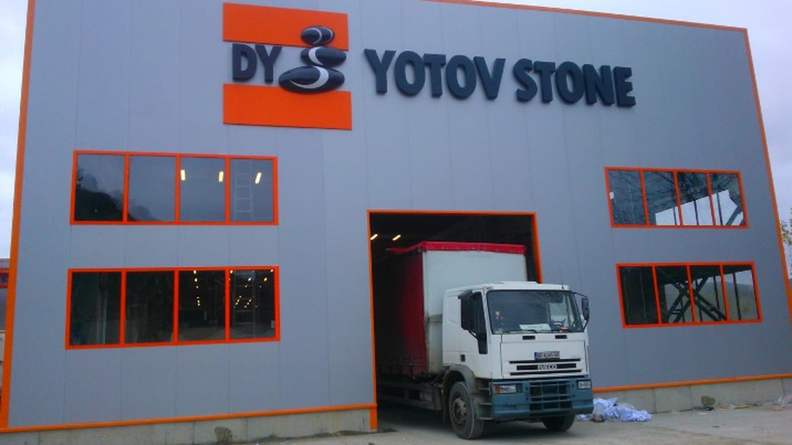 Фасада на сива индустриална сграда с голям черен надпис "Yotov Stone" и камион на входа.