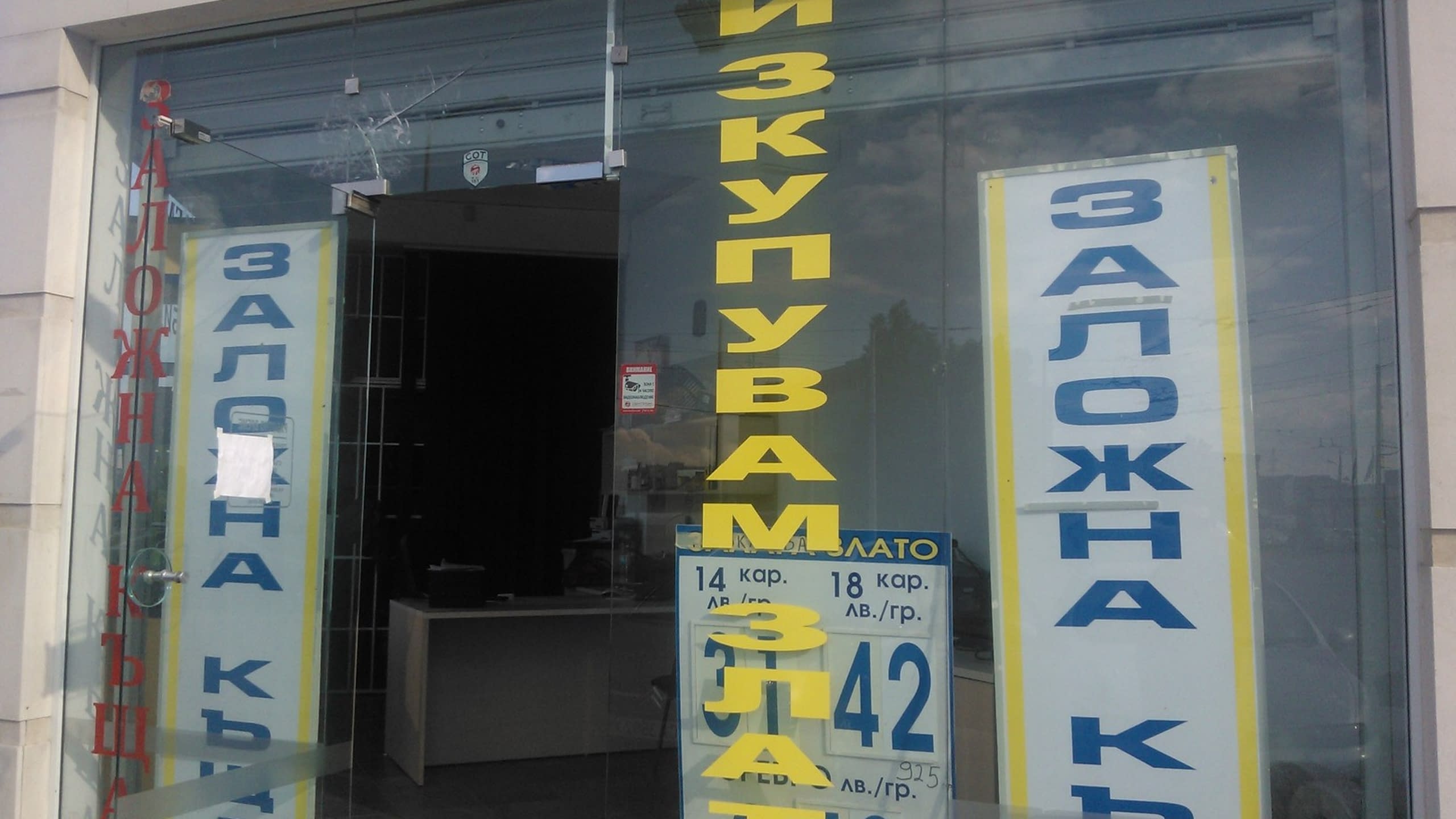 Стъклена врата на заложна къща, облепена с жълти и сини надписи от фолио, информиращи за изкупуване на злато.