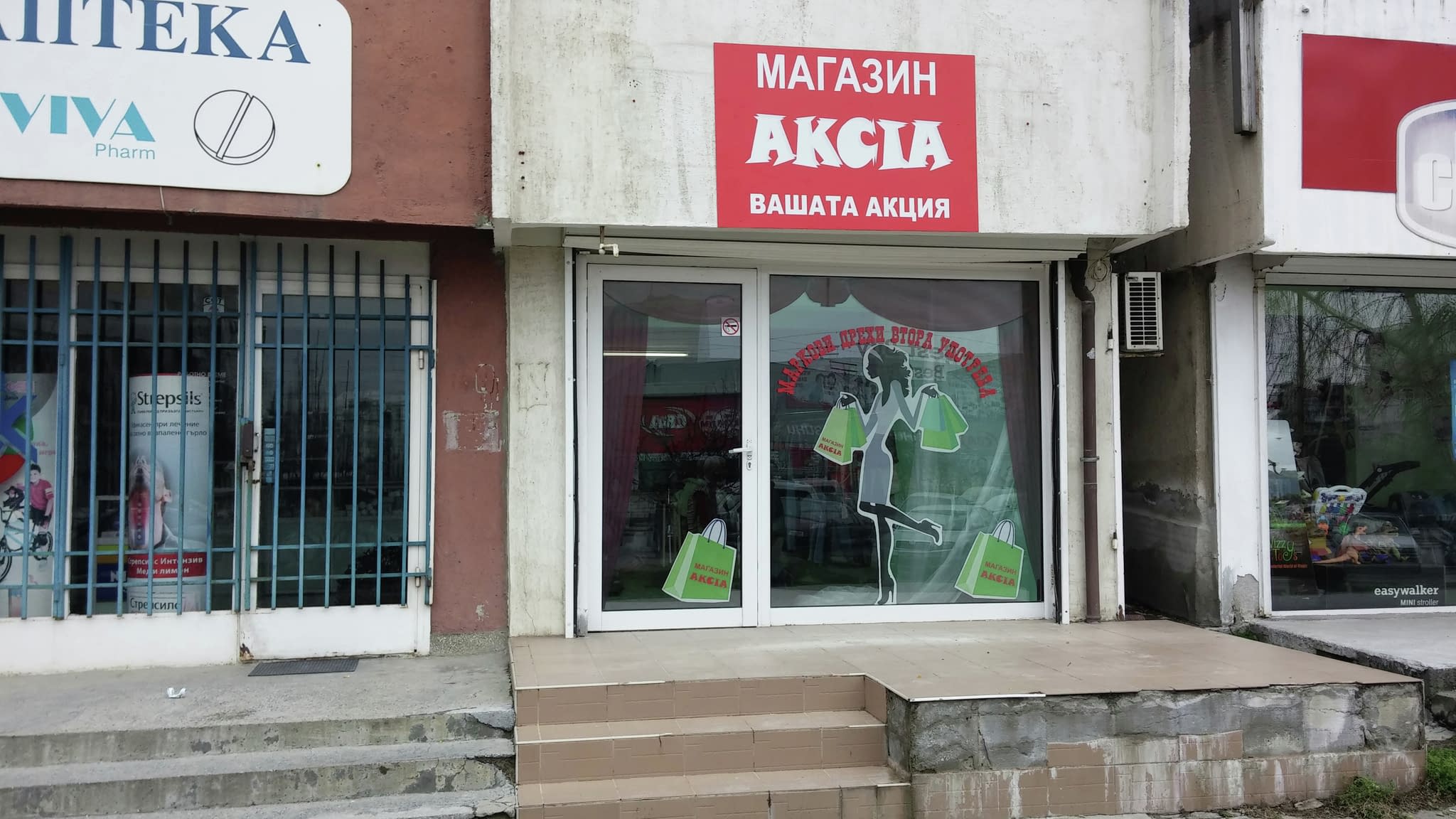 Витрина на ‘Магазин Акция’ със силует на жена с пазарски чанти, изработен от фолио, разположена до аптека във градска среда.