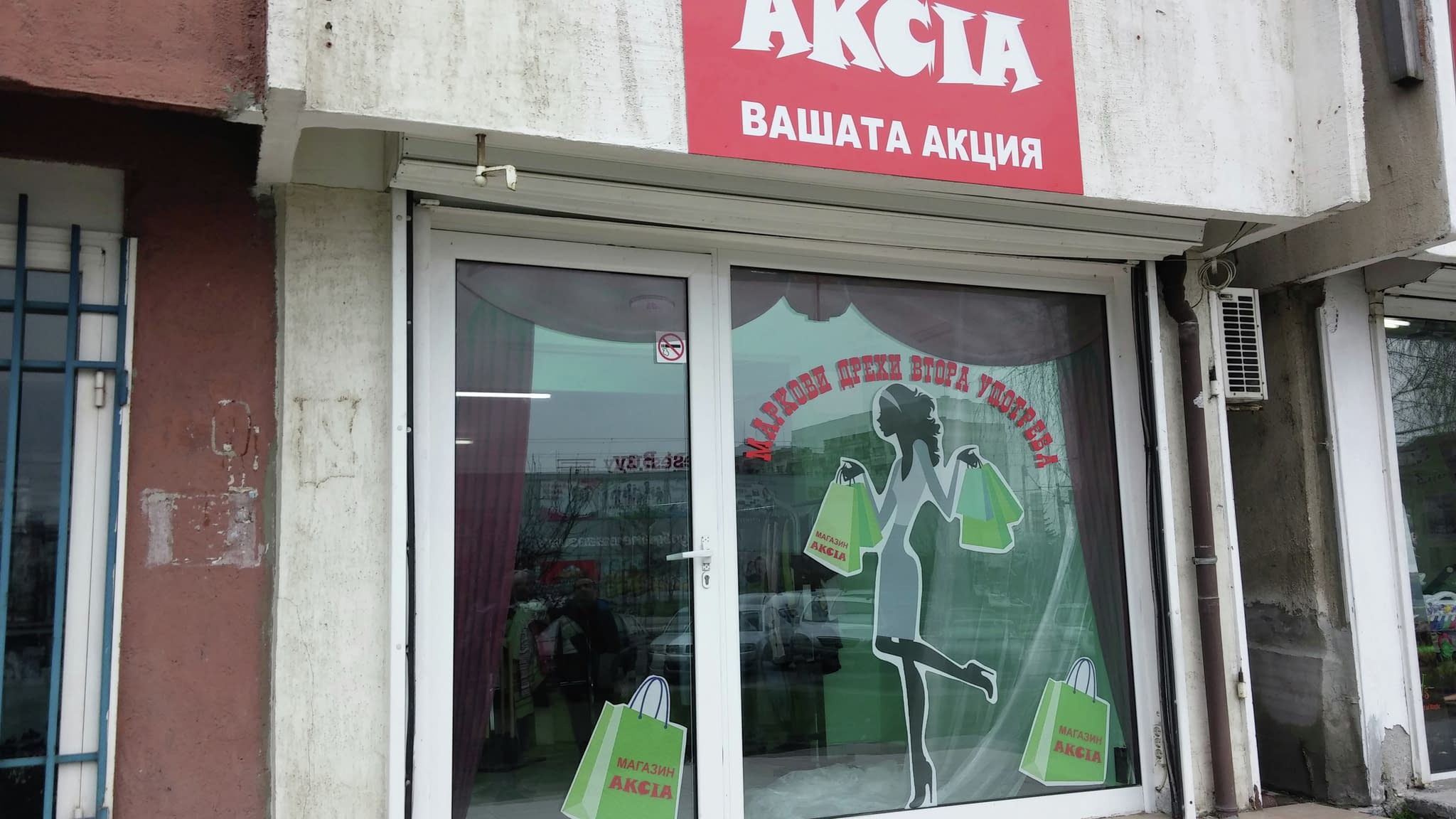 Фасада на магазин ‘Акция’ с червен надпис и витрина, украсена с фолио със силует на жена с пазарски чанти и слоган ‘Маркови стоки, нови усещания’.