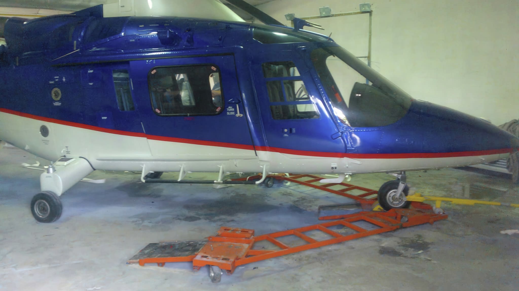 Хеликоптер със син и бял дизайн върху оранжева платформа в хангар.