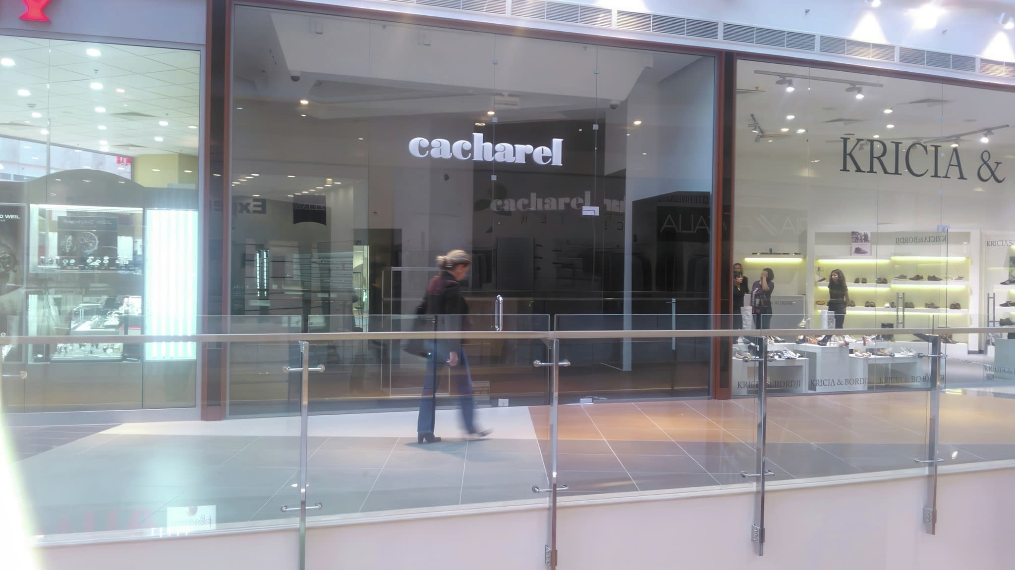 Коридор на търговски център с витрини на магазини Cacharel и Kricia & Co, с преминаващ посетител на преден план.