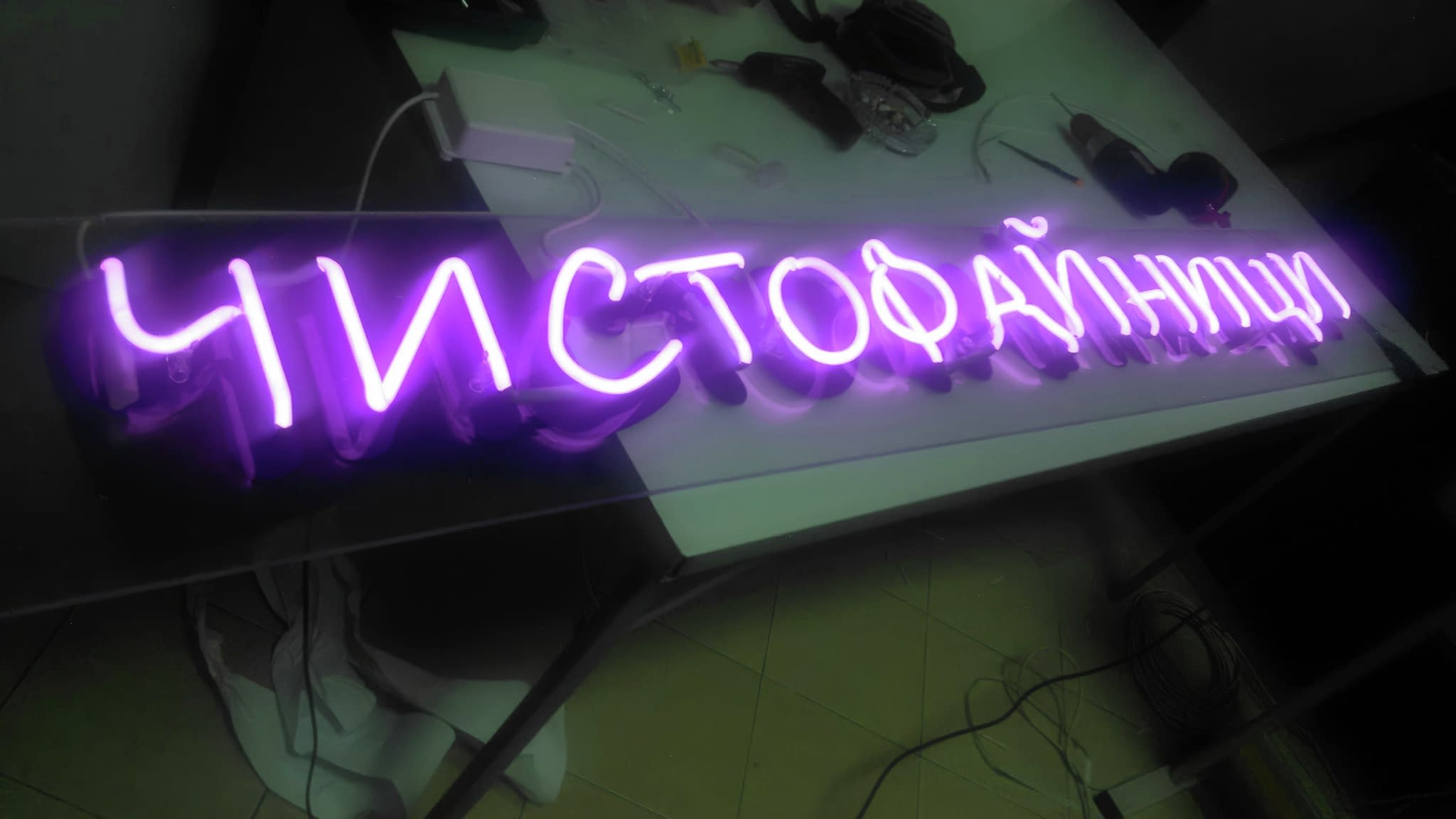 Светещ лилав неонов надпис „ЧИСТОФАЙНИЦИ“, изложен на работна маса в производствено ателие.