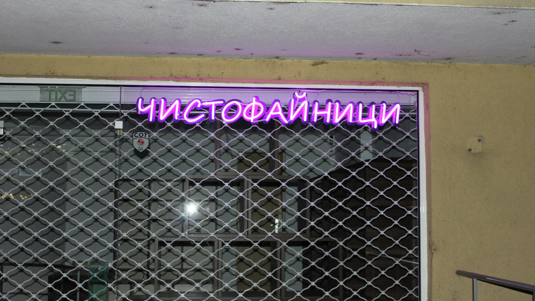 Фасада на затворен магазин с лилава неонова табела „ЧИСТОФАЙНИЦИ“, монтирана над решетъчна охранителна врата.