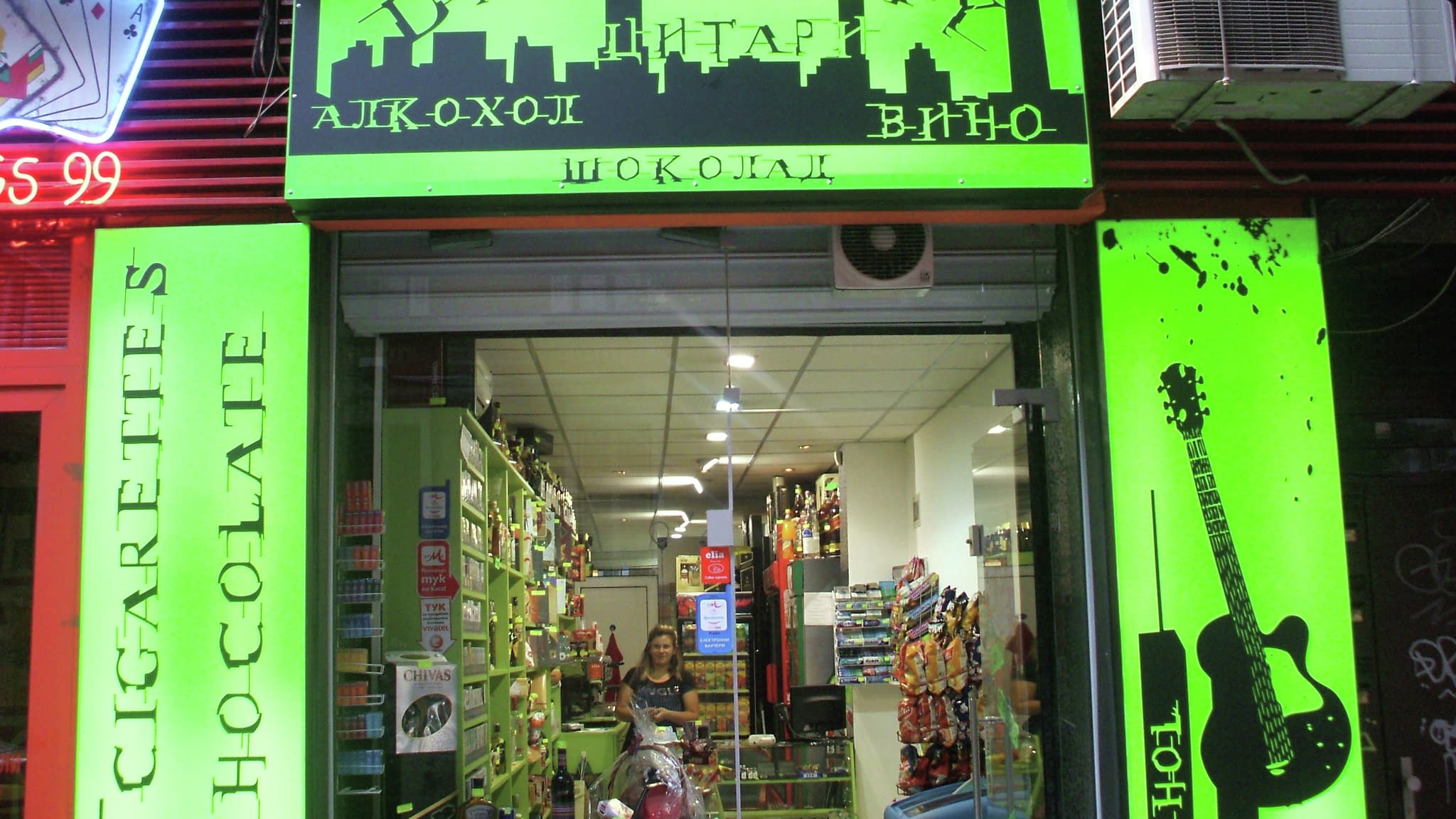 Фасада на магазин „Dark City“ в София с ярка зелена светеща реклама, включваща силует на градски пейзаж, надписи за цигари, шоколад, алкохол и вино.
