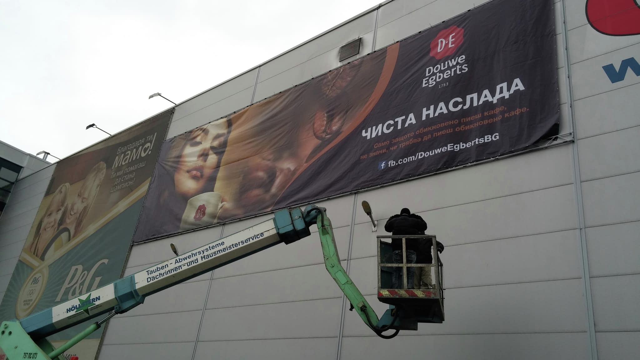 Работник в автовишка монтира рекламен банер за кафе Douwe Egberts на фасада, с изображение на жена, която държи чаша кафе.
