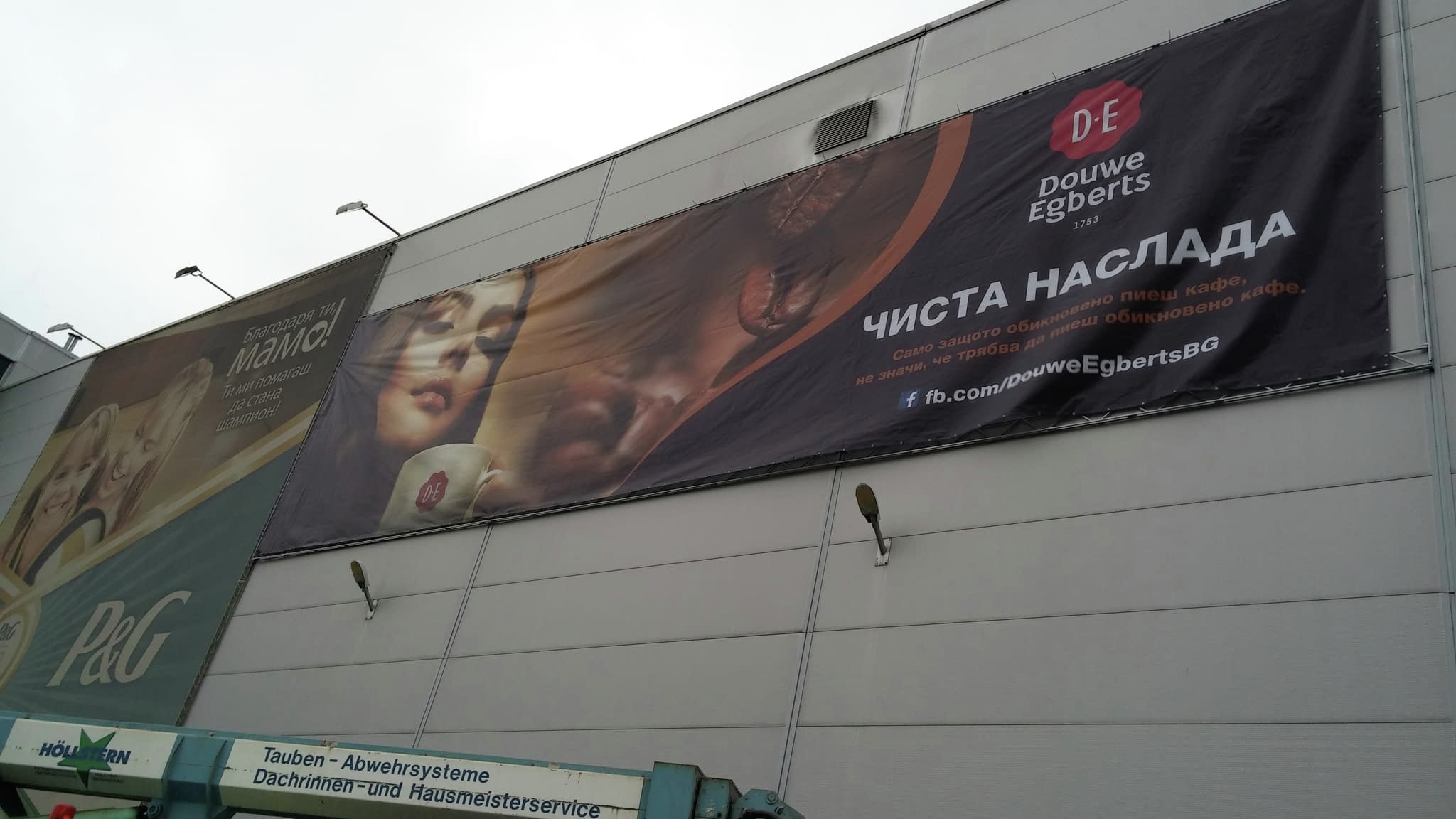 Рекламен банер за кафе Douwe Egberts с надпис „Чиста наслада“ и линк към Facebook страница, окачен върху фасадата на сграда.