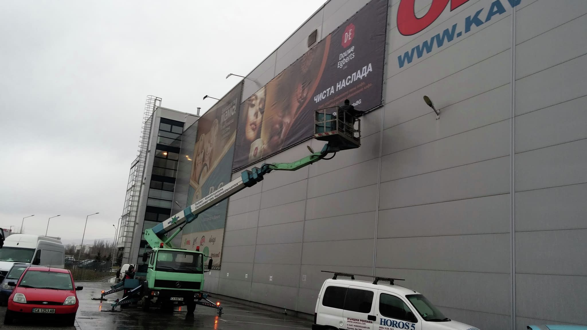 Рекламен транспарант за кафе Douwe Egberts, монтиран на голяма фасада с помощта на автовишка, показващ слоган „Чиста наслада“ и чаша кафе.