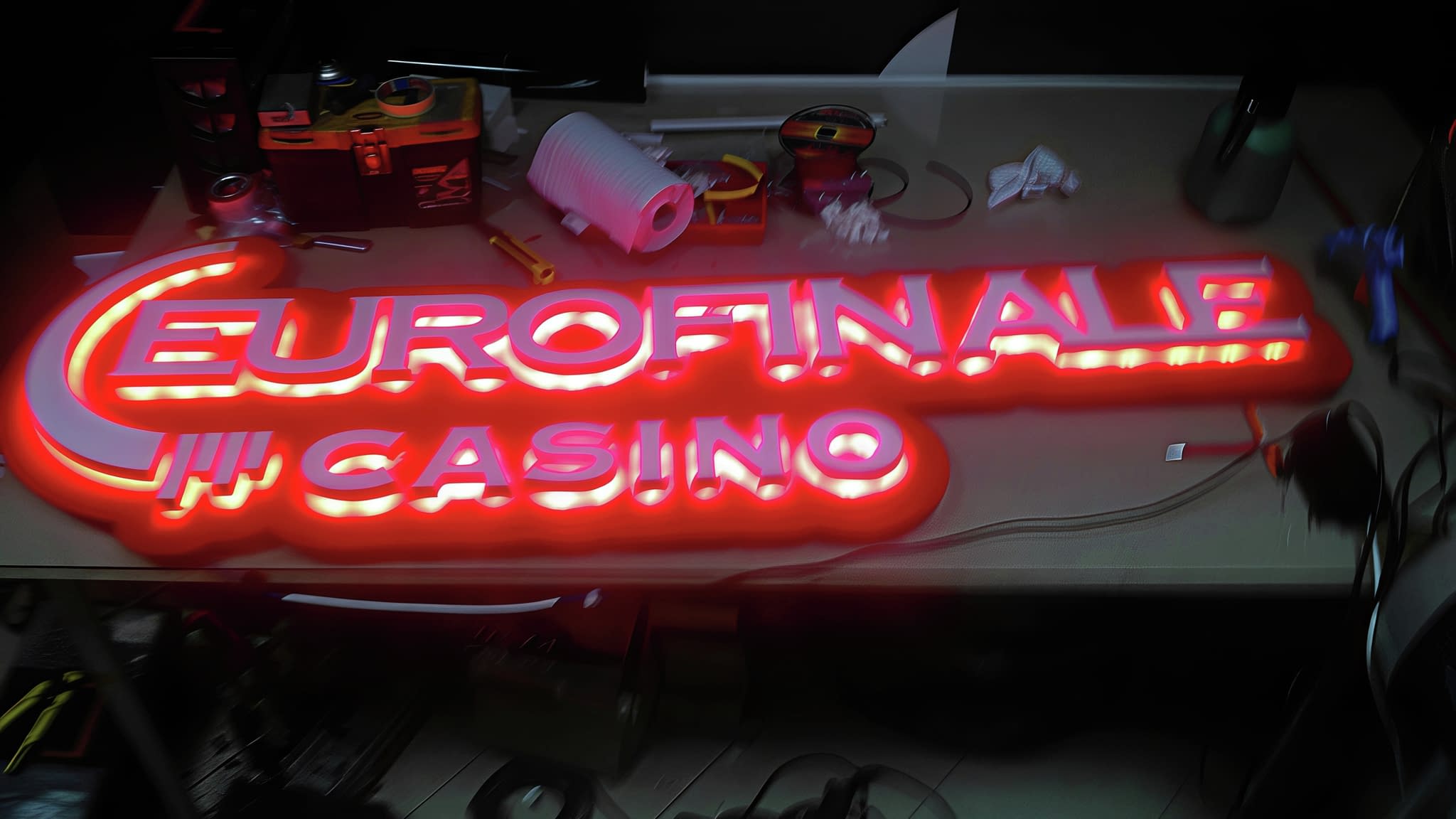 LED табела 'EUROFINALE CASINO' с червен неонов ефект, поставена върху работна маса със строителни инструменти и материали.