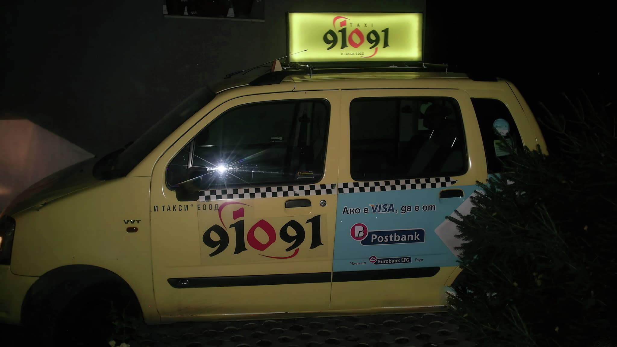 Жълто такси с брандирана реклама на номер „91091“ и логото на И Такси ЕООД, със странична реклама за плащане с VISA, Postbank и Eurobank, заснето през нощта.