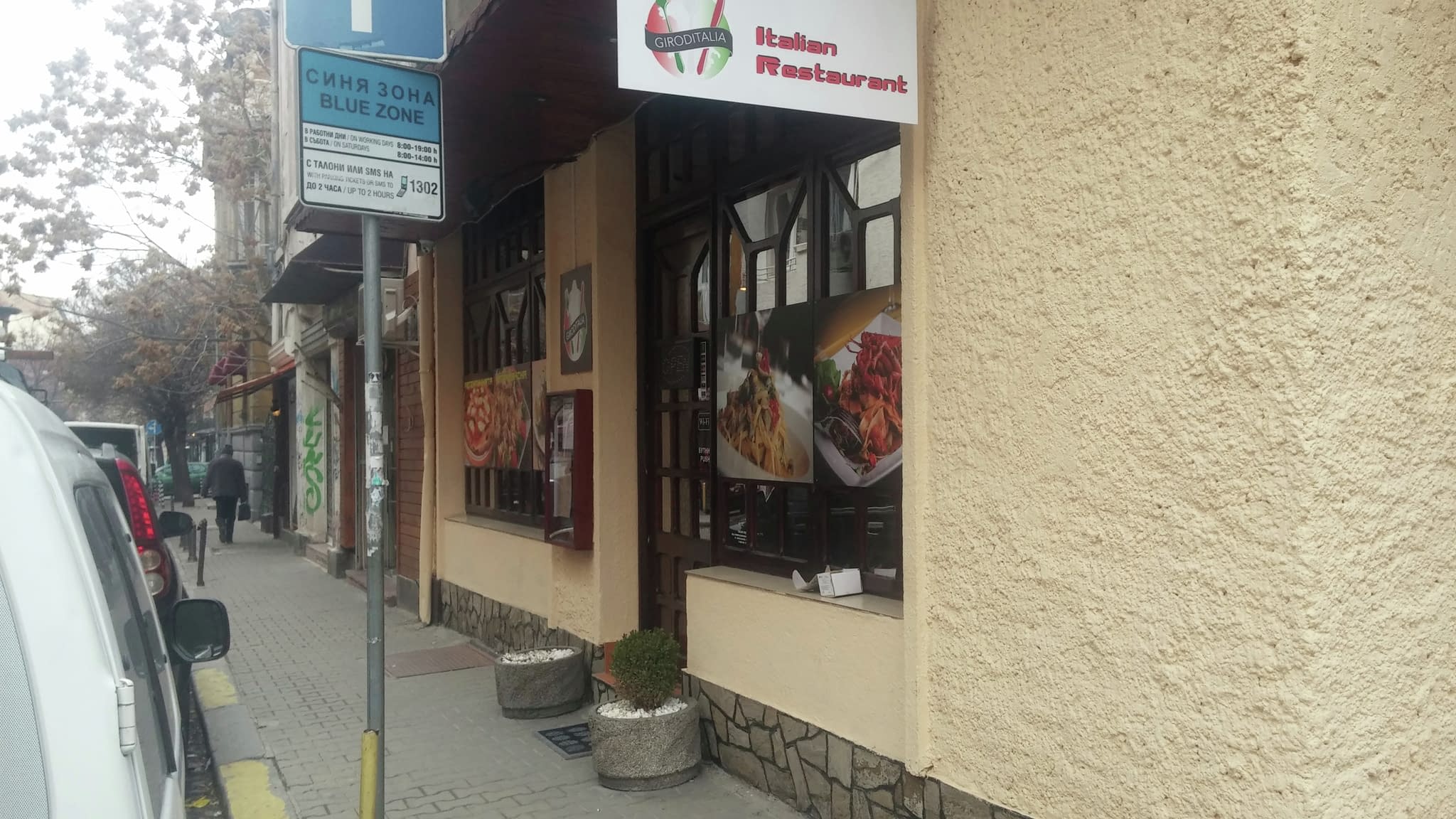 Екстериор на италиански ресторант с двуезична табела на български и английски, витрина с изображения на пица и паста, както и знак за синя зона паркинг наблизо.