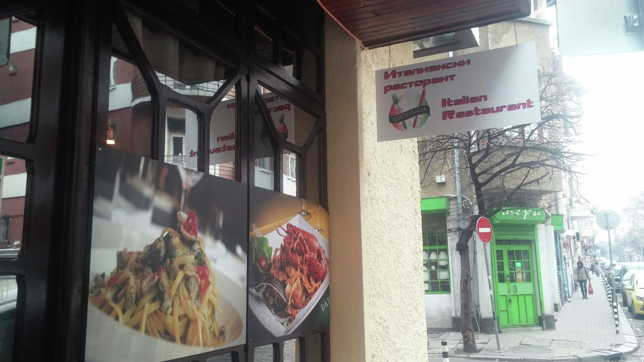 Външен изглед на италиански ресторант с двуезична табела ‘Italian Restaurant’ и рекламни снимки на витрината, представящи паста и месни ястия.