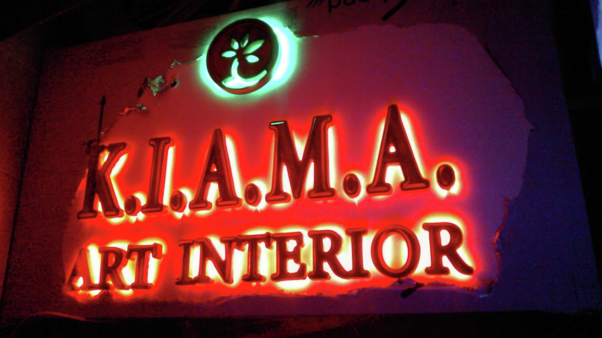 Рекламна табела с червено неоново осветление, надпис ‘K.I.A.M.A. Art Interior’ и стилизирано лого на фона, подходяща за външна реклама.