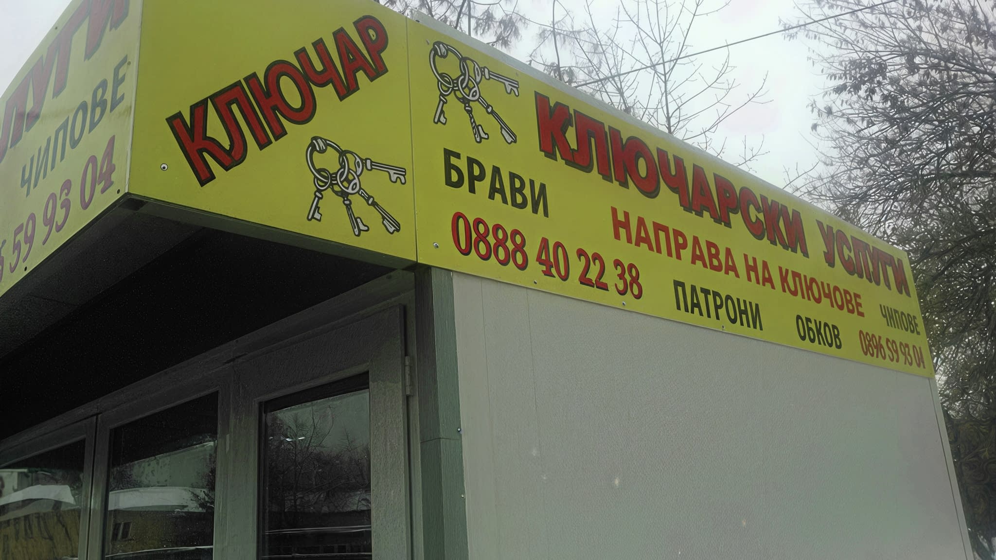 Информационна табела за ключар с надпис „Ключарски услуги“ и жълт фон, рекламиращ услуги като направа на ключове, патрони и чипове.